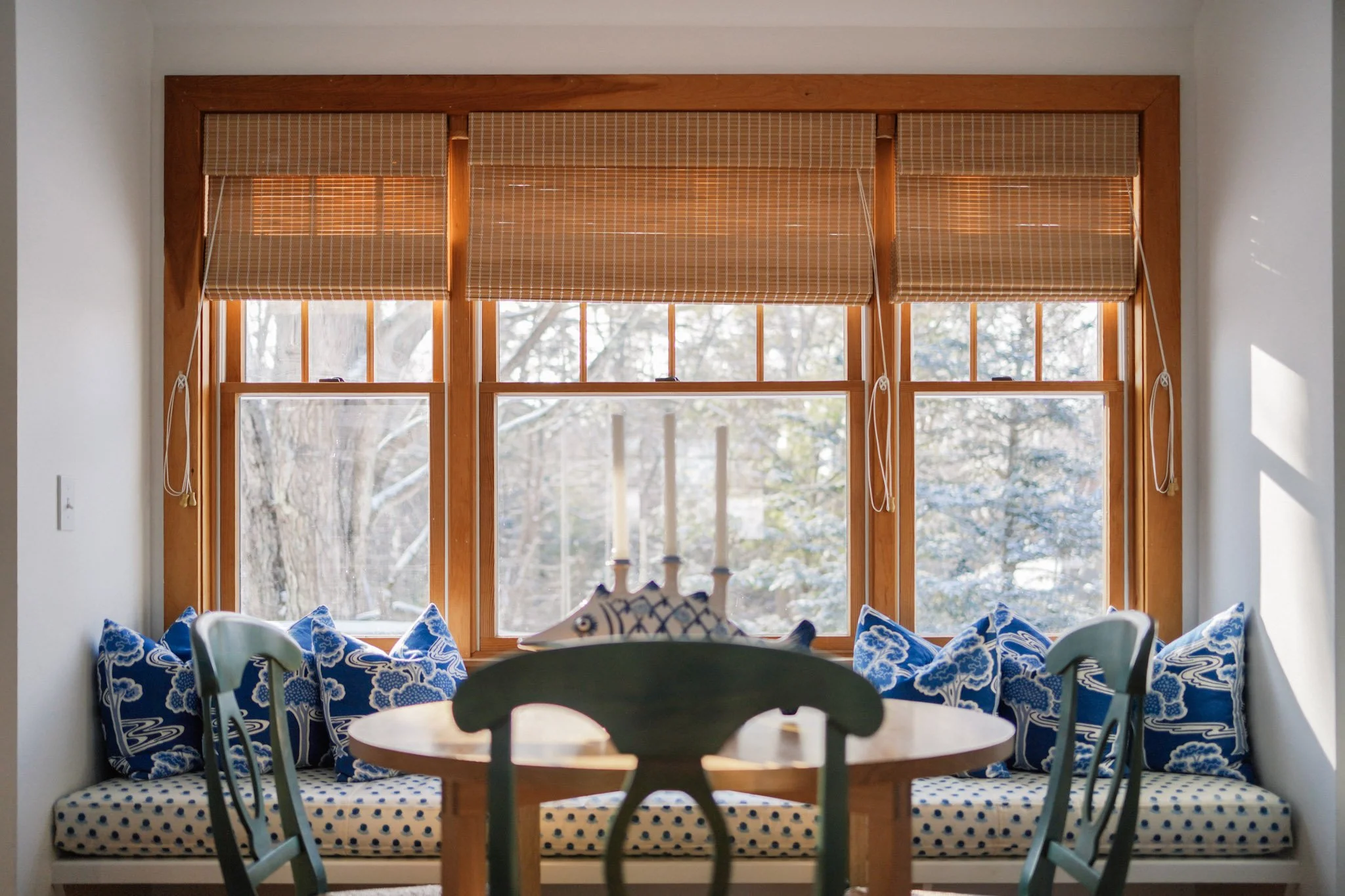 A built-in window seat with blue and white pillows, bamboo blinds, and a dining table in the foreground