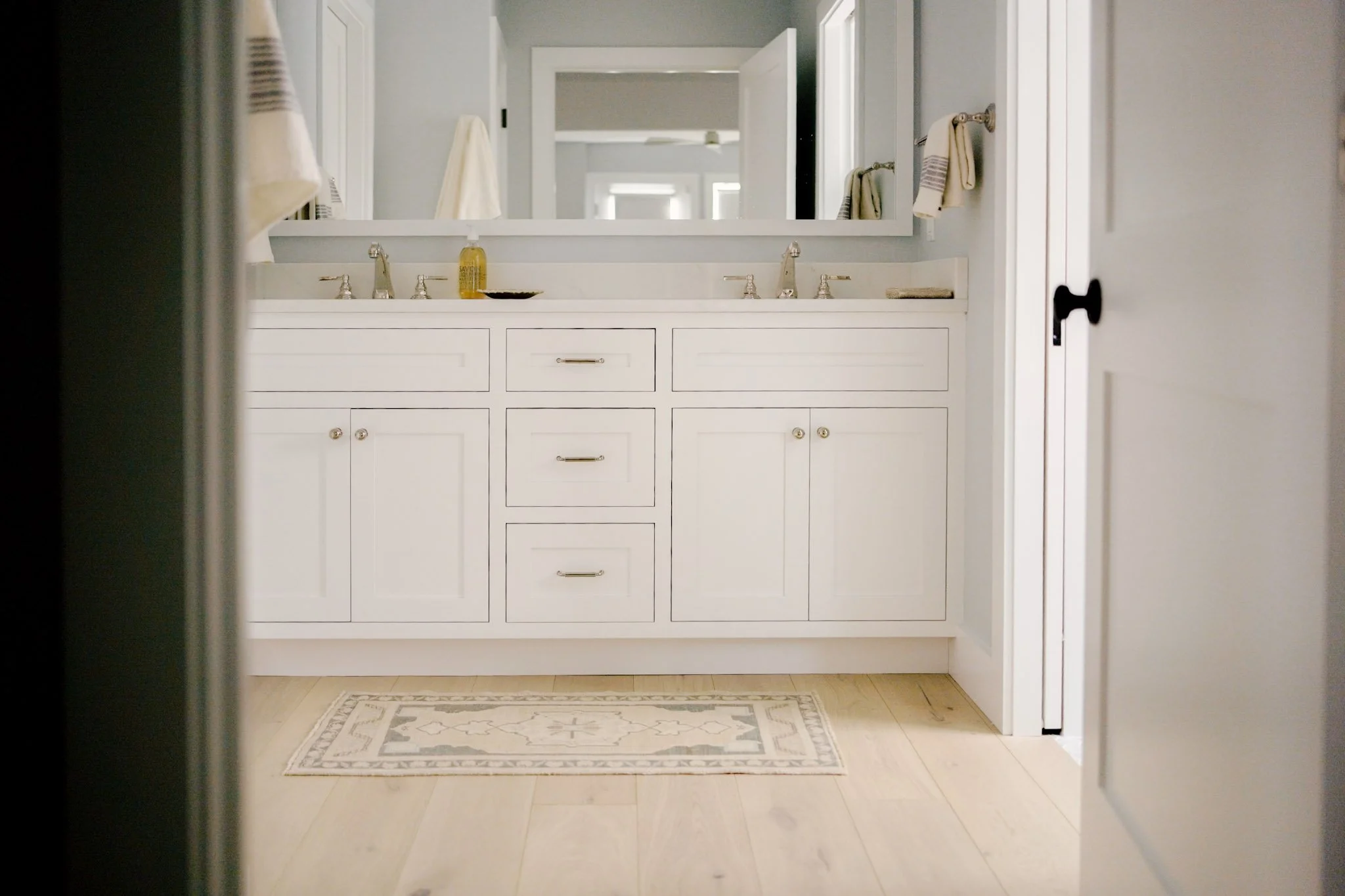 A double vanity bathroom with two sinks, a large mirror, and a light wood floor with a decorative rug
