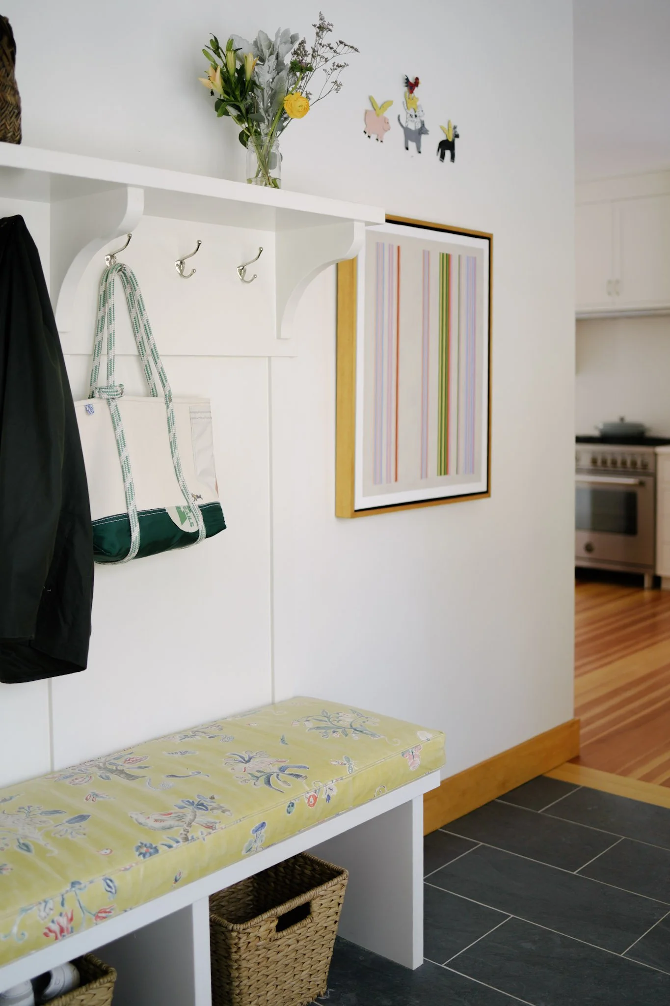 A hallway with a white painted hall tree, floral bench cushion, woven baskets, and a piece of framed striped artwork on black tile flooring