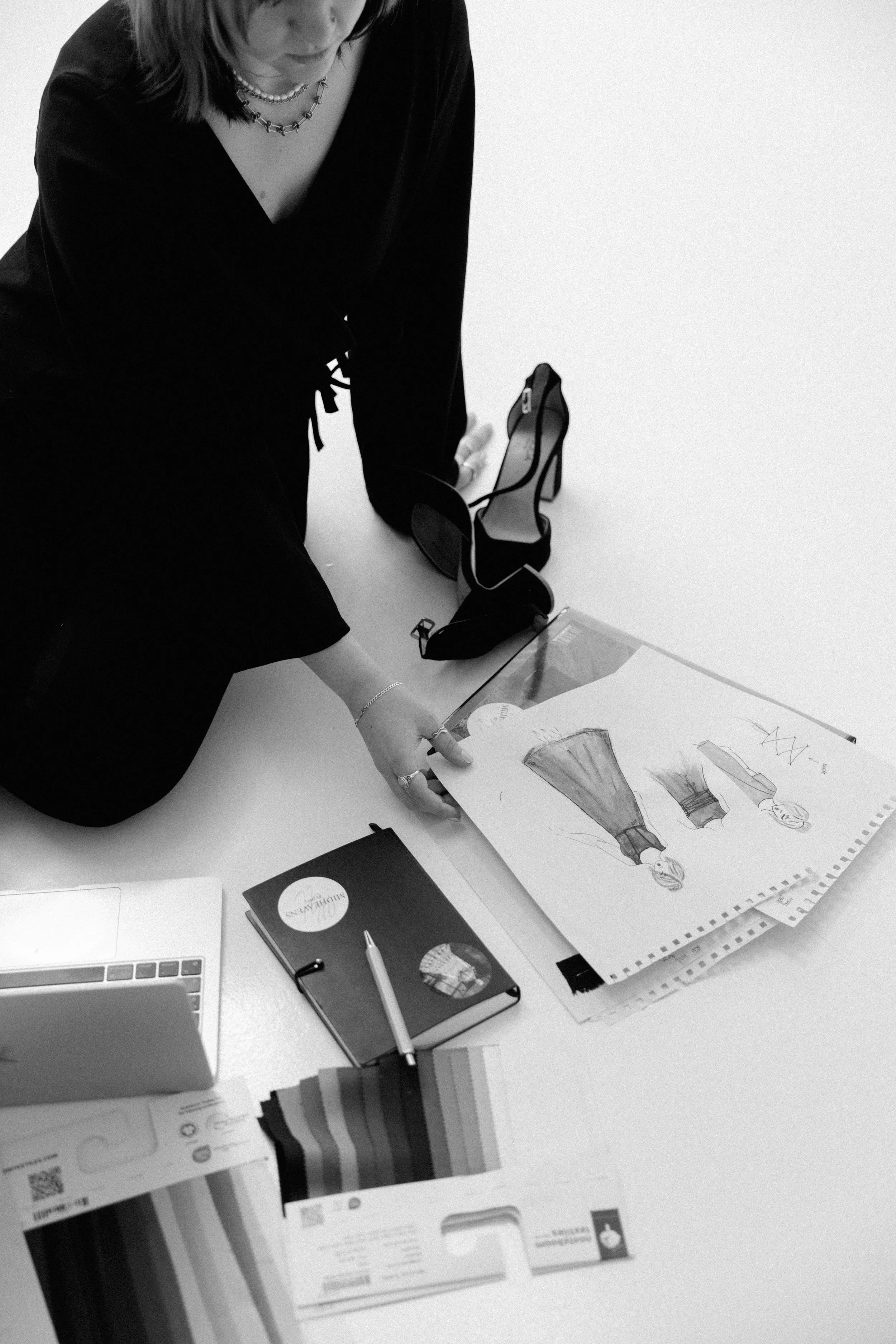 A woman sitting at a desk looking at and sketching fashion designs on paper. The desk has various documents, a closed laptop, and a pen.