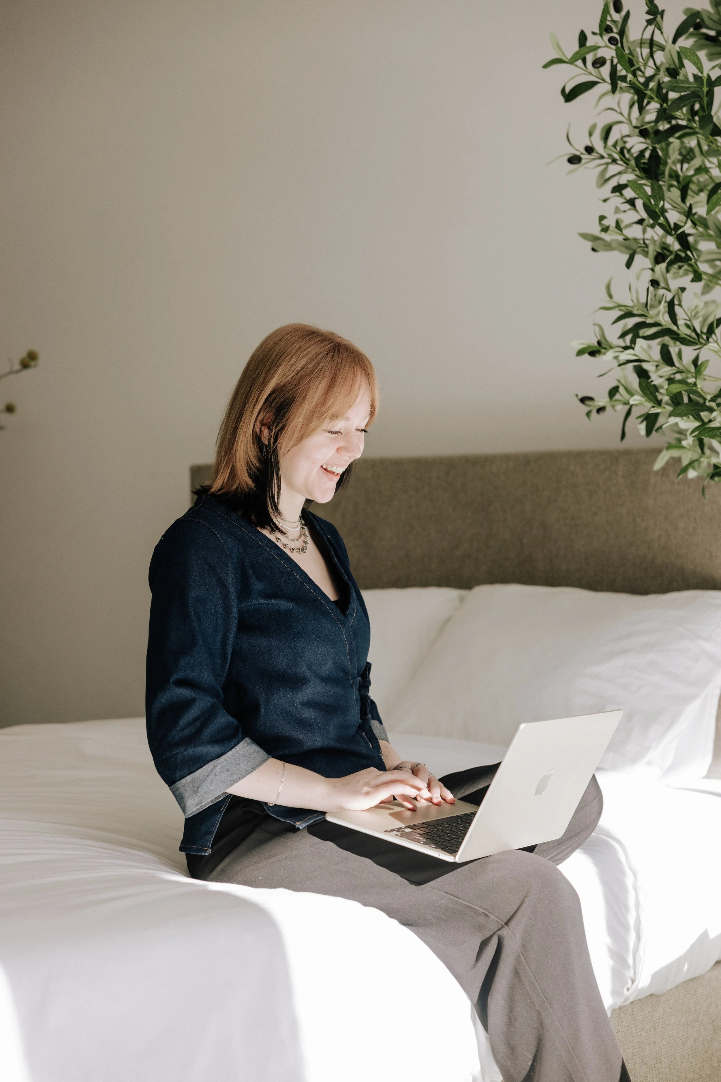woman sitting on a bed using a silver MacBook laptop, smiling, with a green plant and a beige headboard in the background.