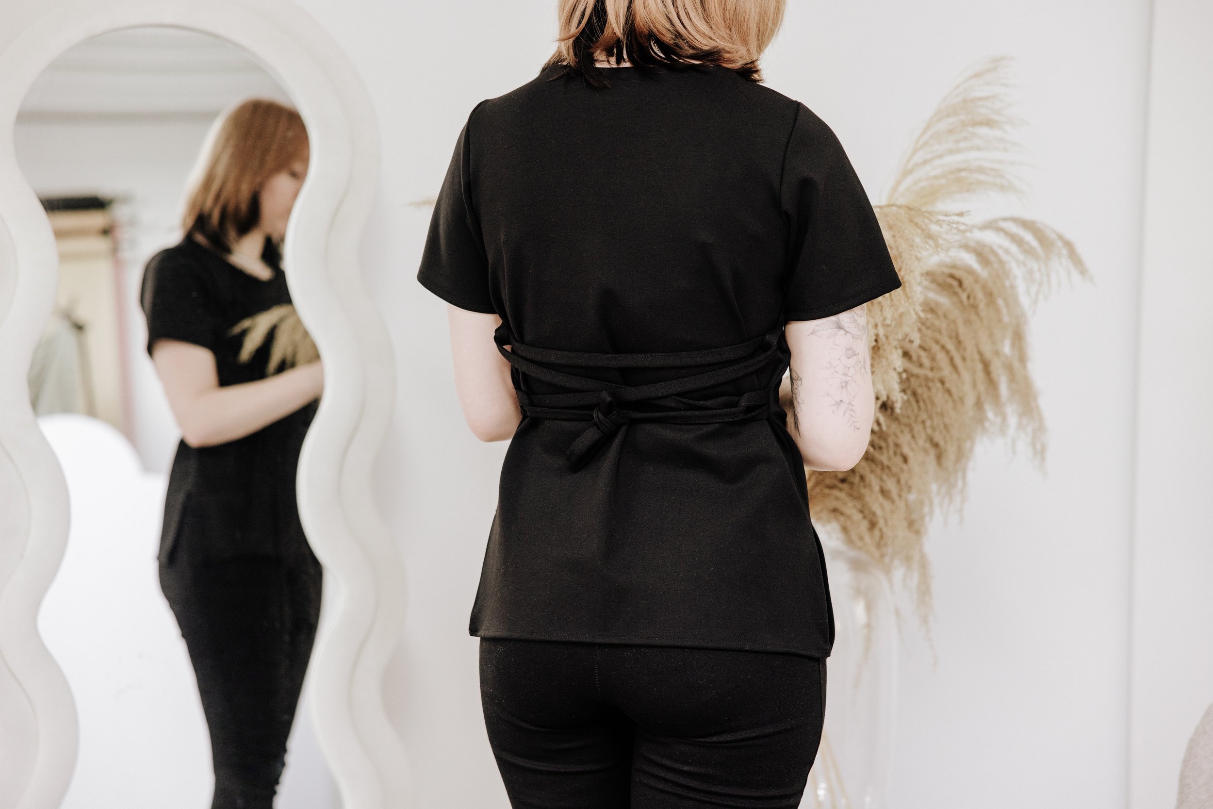 Woman with shoulder-length hair is standing in front of a mirror, wearing a black dress with a tied waist, and looking at her reflection.