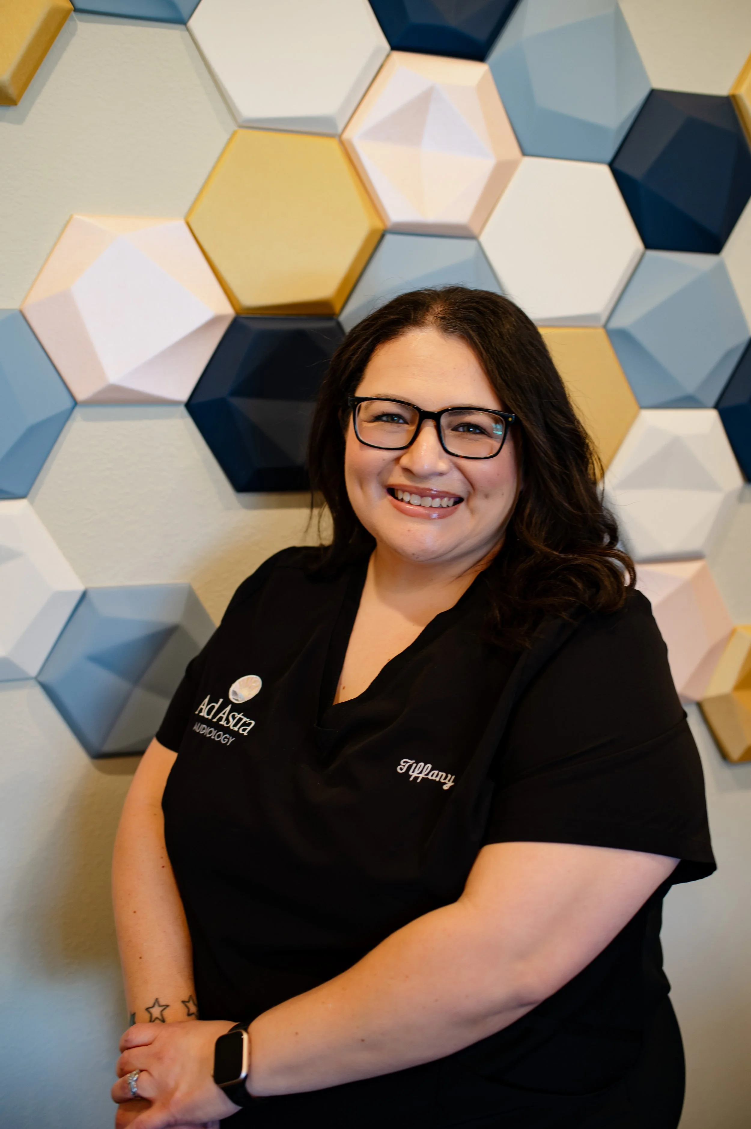 A smiling woman with dark hair and glasses sitting against a wall with colorful 3D hexagonal wall decor. She is wearing a black top with embroidery and a smartwatch on her left wrist.