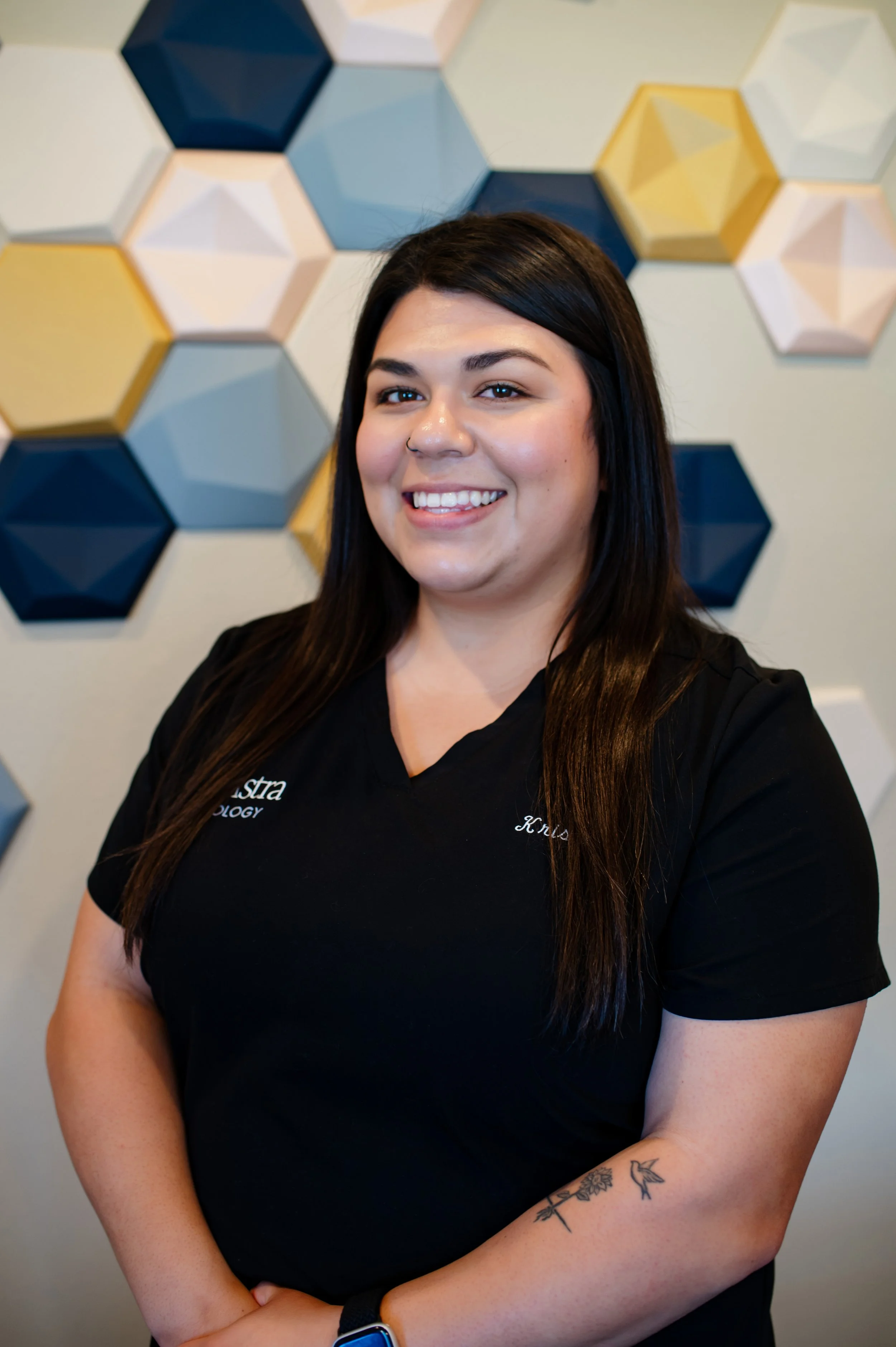 A woman with dark hair smiling, wearing a black shirt with her arms crossed, standing in front of a wall decorated with colorful hexagonal wall art.