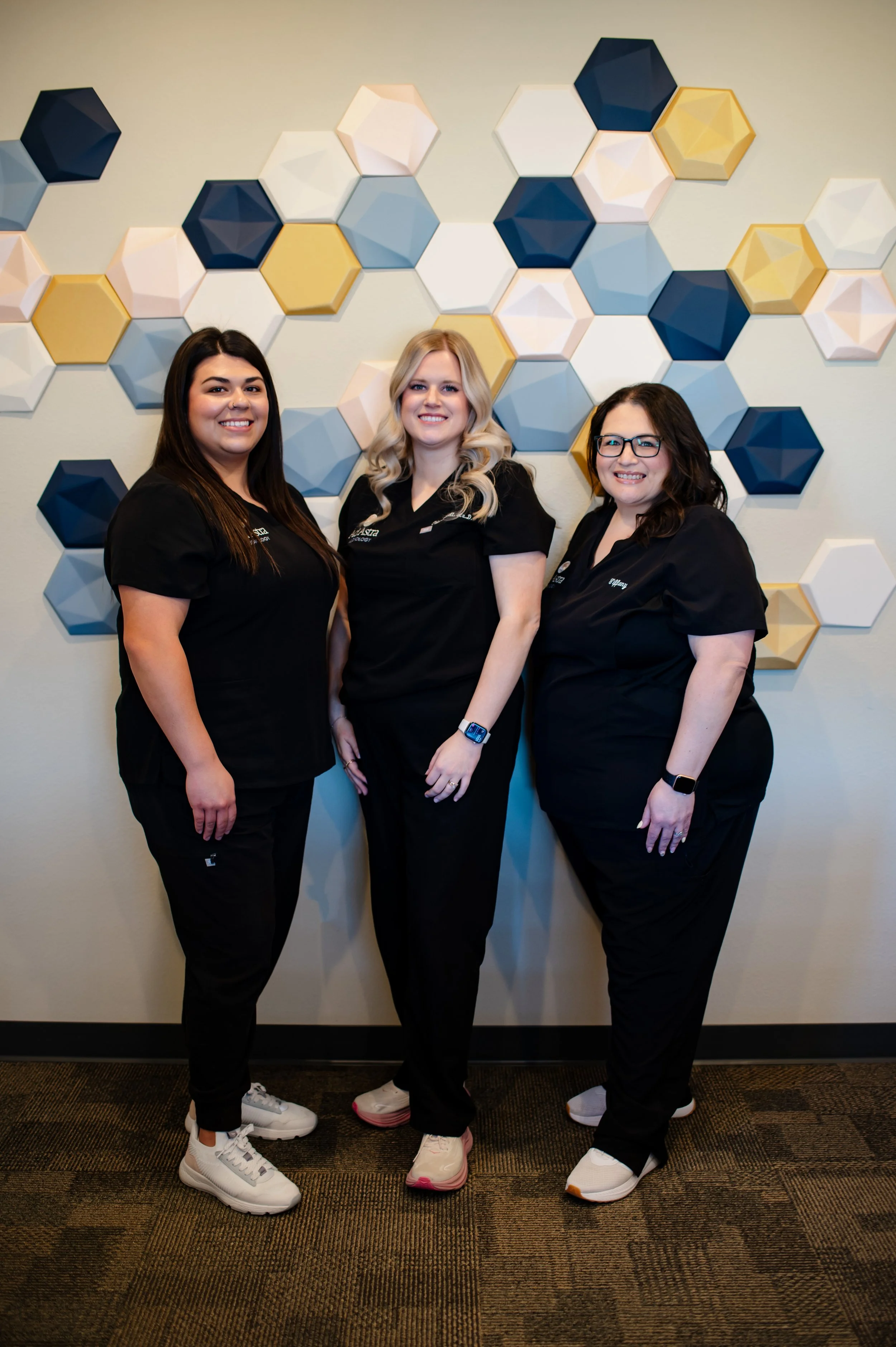 Three women dressed in black scrubs standing in front of a wall decorated with hexagonal 3D wall art in shades of yellow, white, blue, and dark blue. They are smiling and posing for the photo.