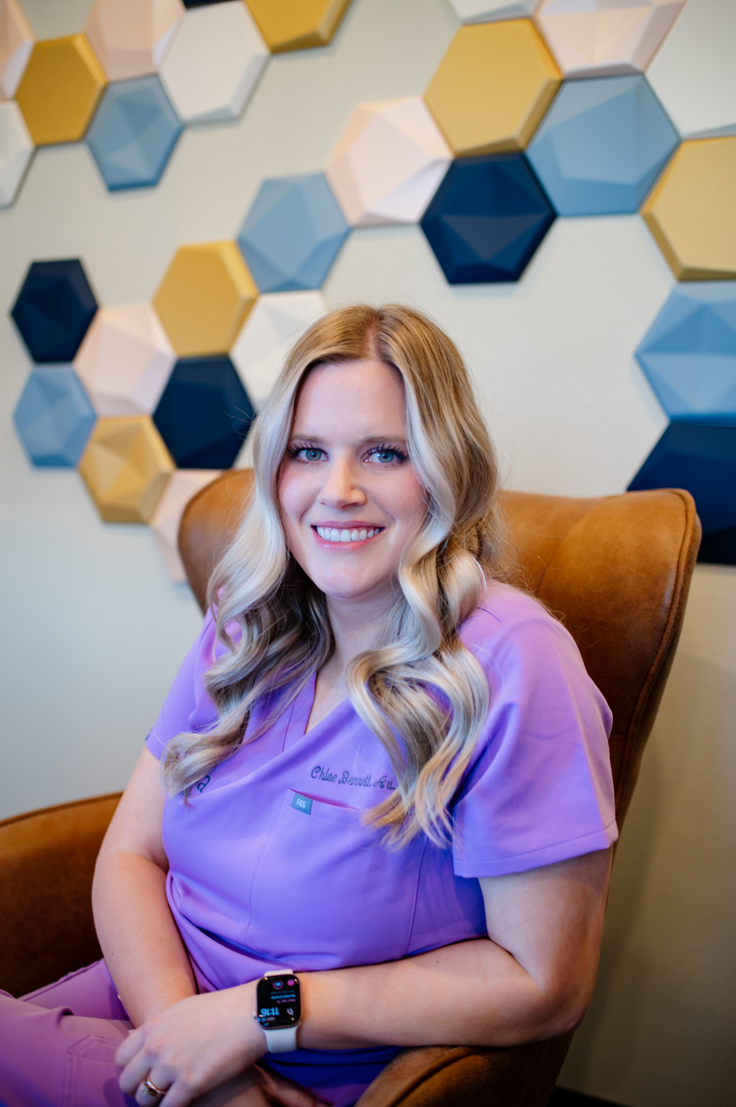 A woman wearing purple medical scrubs, with long wavy blonde hair and blue eyes, smiling while sitting in a brown armchair. She is wearing a smartwatch on her left wrist and a ring on her right hand. Behind her, there is a wall decorated with multicolored hexagonal 3D art pieces in shades of blue, white, yellow, and gold.