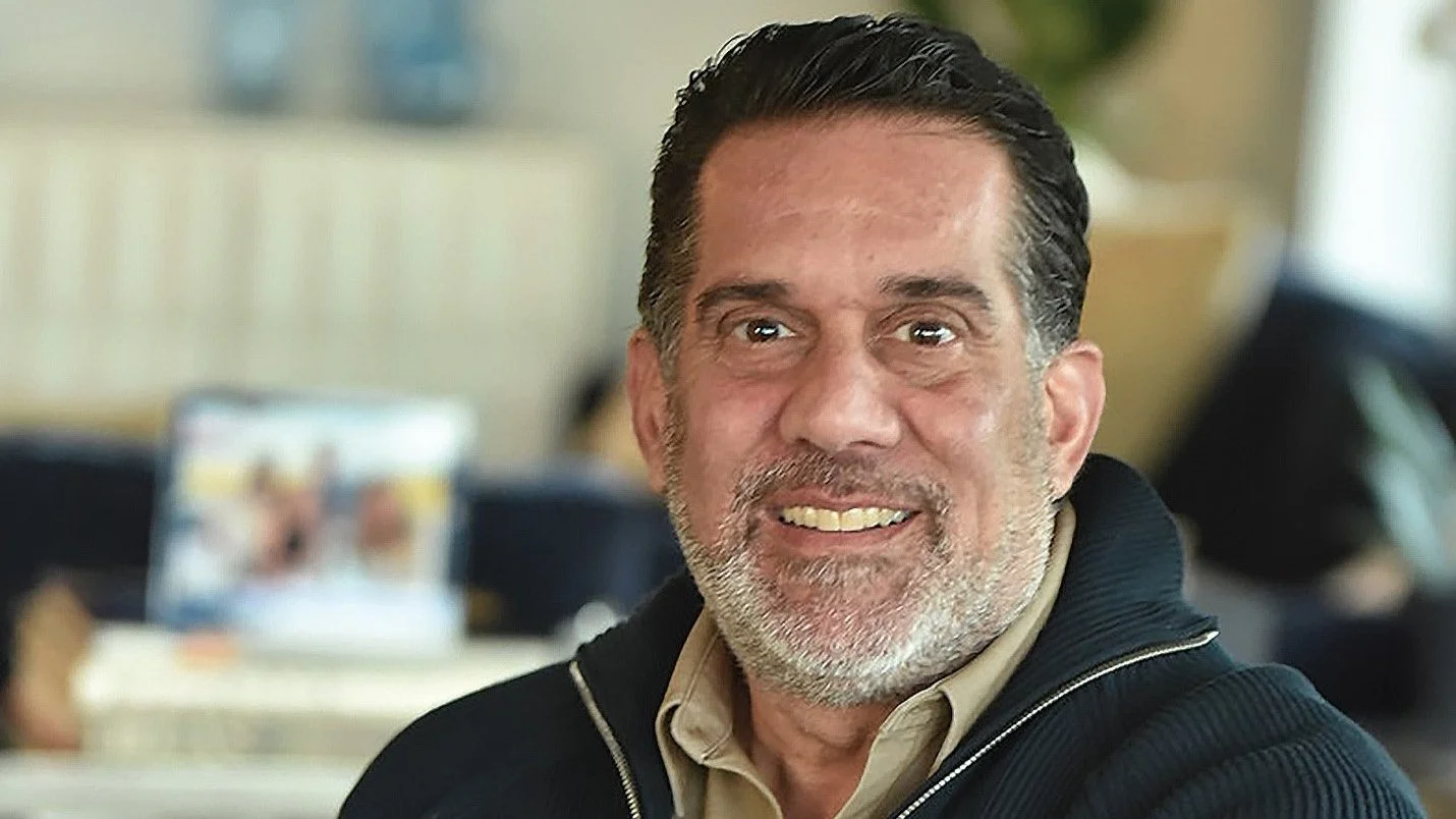 Smiling man with a beard and dark hair, wearing a dark jacket in an indoor setting.