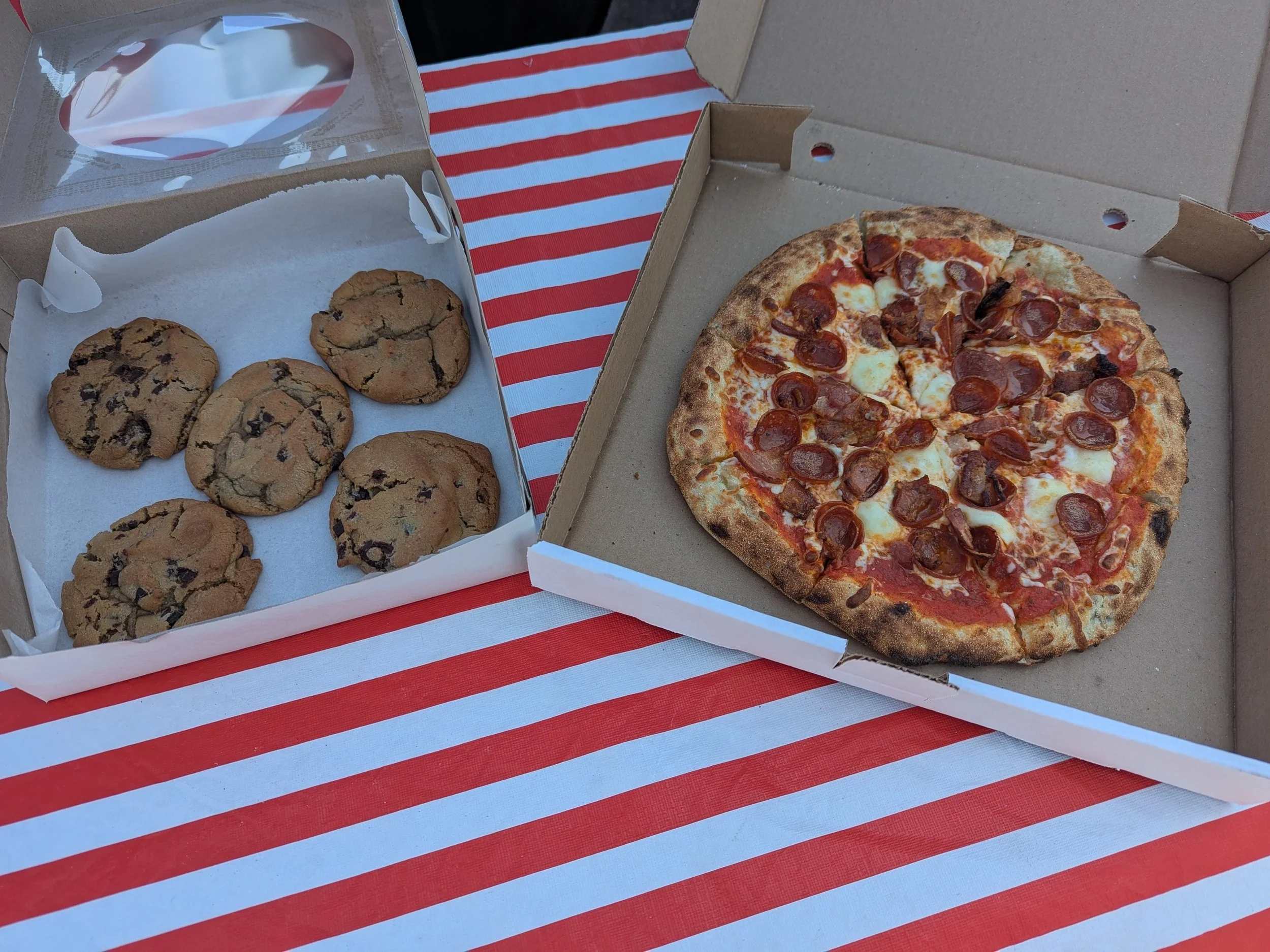 A pizza with pepperoni and cheese in a cardboard box and a box of six chocolate chip cookies on a red and white striped tablecloth.