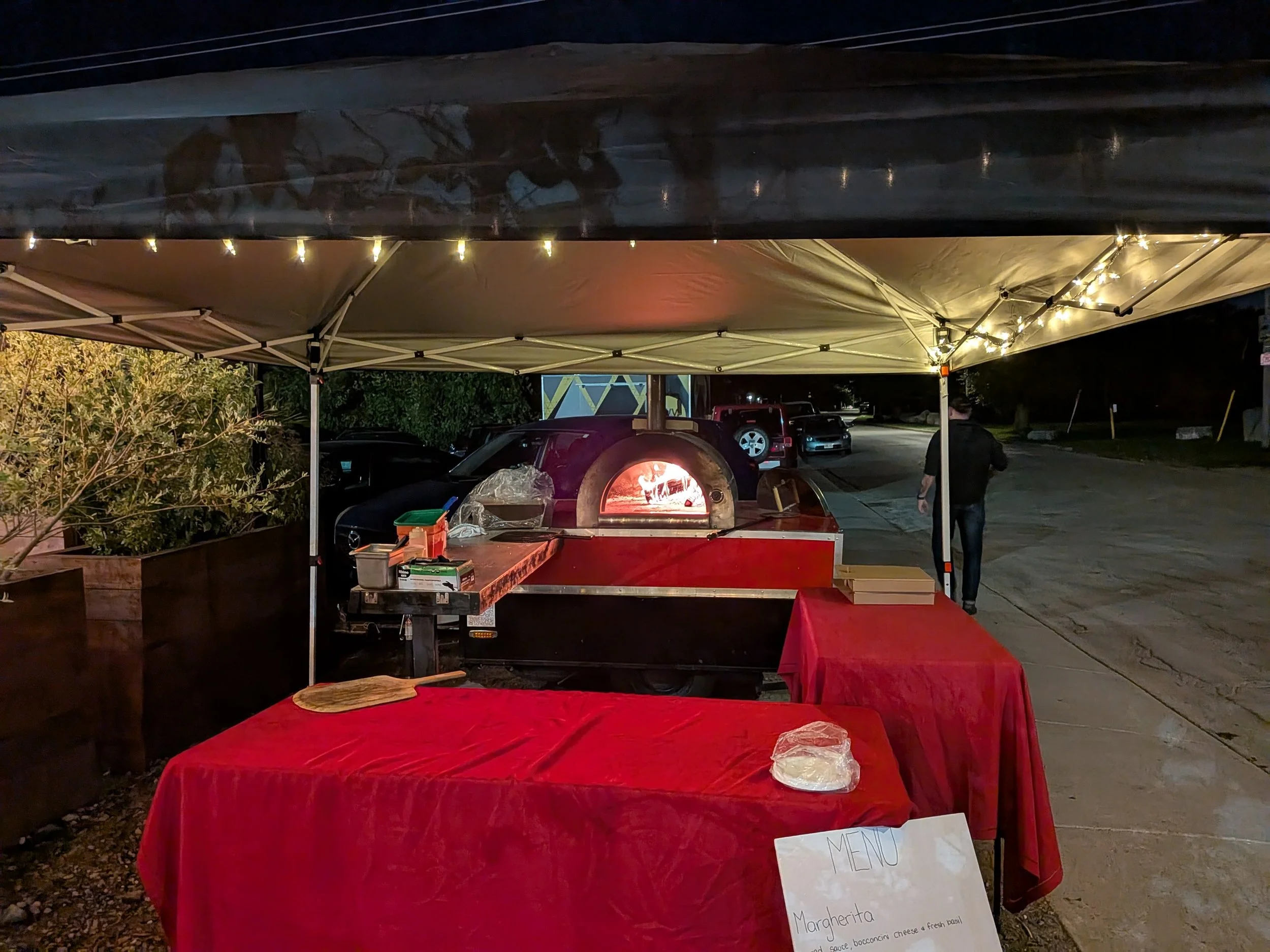 Nighttime outdoor pizza stand with a wood-fired pizza oven, red tables covered with red cloth, and a handwritten menu with Margherita pizza description.