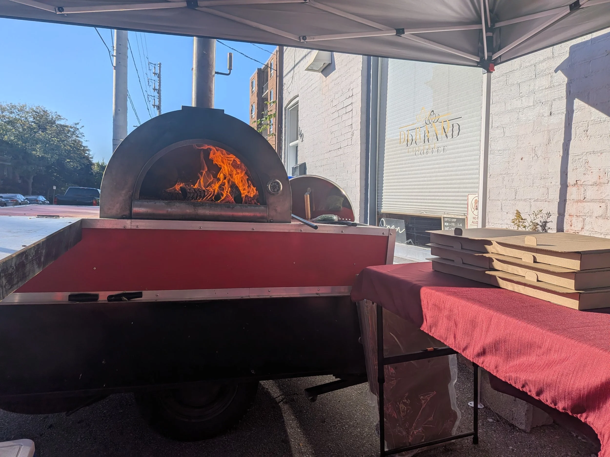 A pizza oven with a visible fire inside, situated on a cart next to a table covered with brown pizza boxes on a red cloth, under an outdoor tent outside a building with a sign that reads 'Dough and Co.'