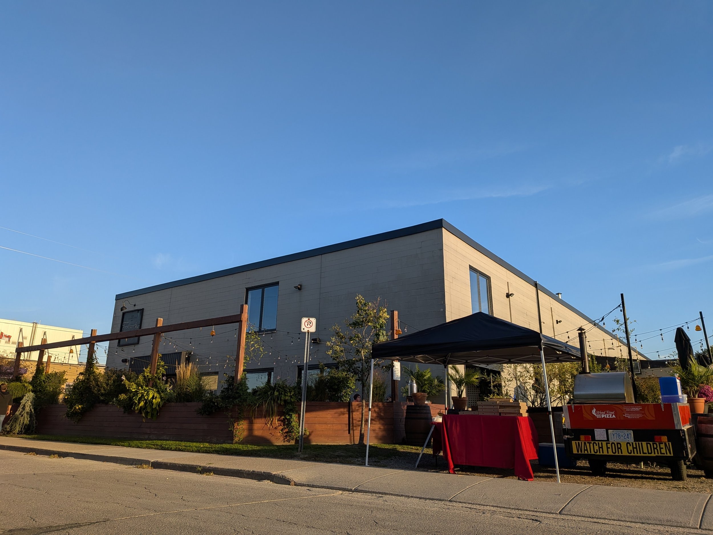 Modern two-story building with large windows, outdoor string lights, and a small vendor stand with a sign that reads 'WATCH FOR CHILDREN' in the foreground, under a black canopy, on a paved street during the day.
