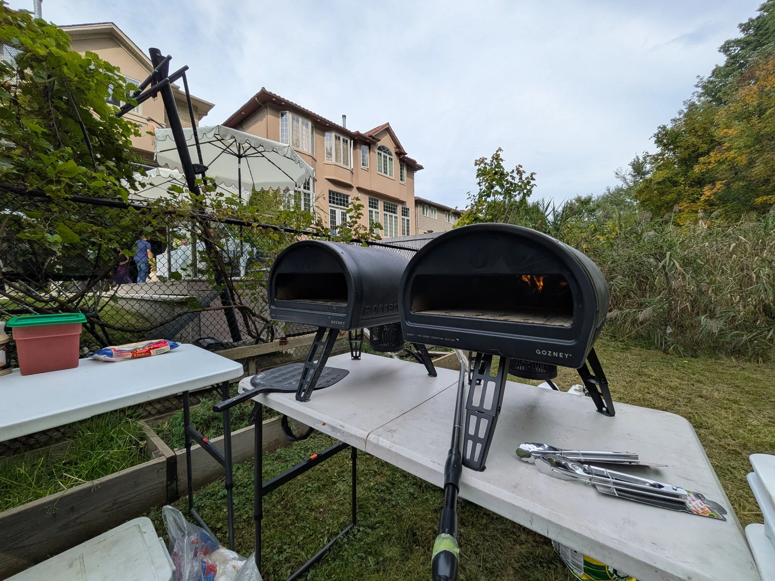 Two black pizza ovens on a white table outdoors, surrounded by grass, plants, and residential buildings, with tongs and kitchen tools nearby.