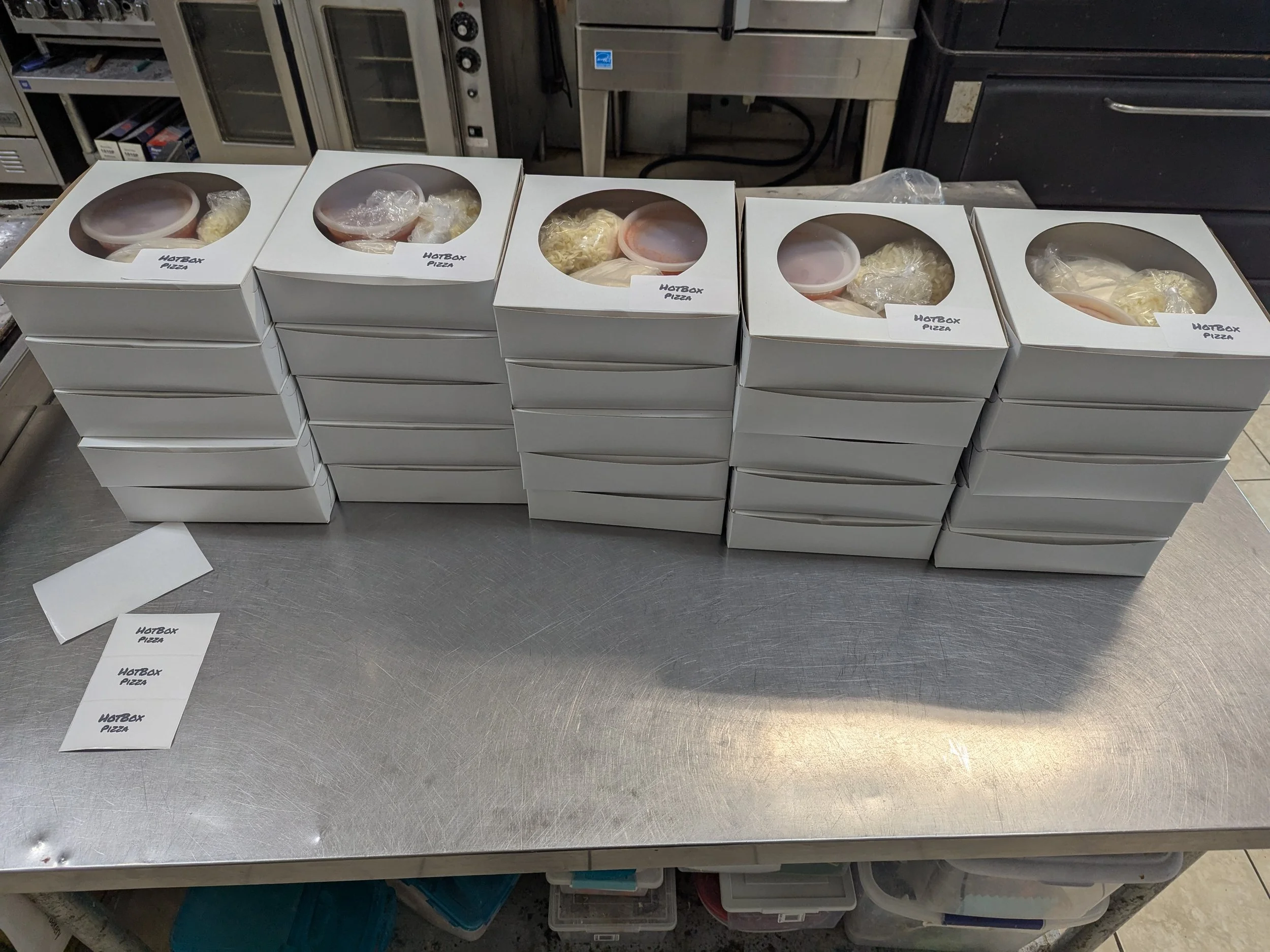 Several white pizza boxes labeled 'HOTBOX PIZZA' stacked on a stainless steel table, with some open showing plastic-wrapped cheese and other ingredients inside.