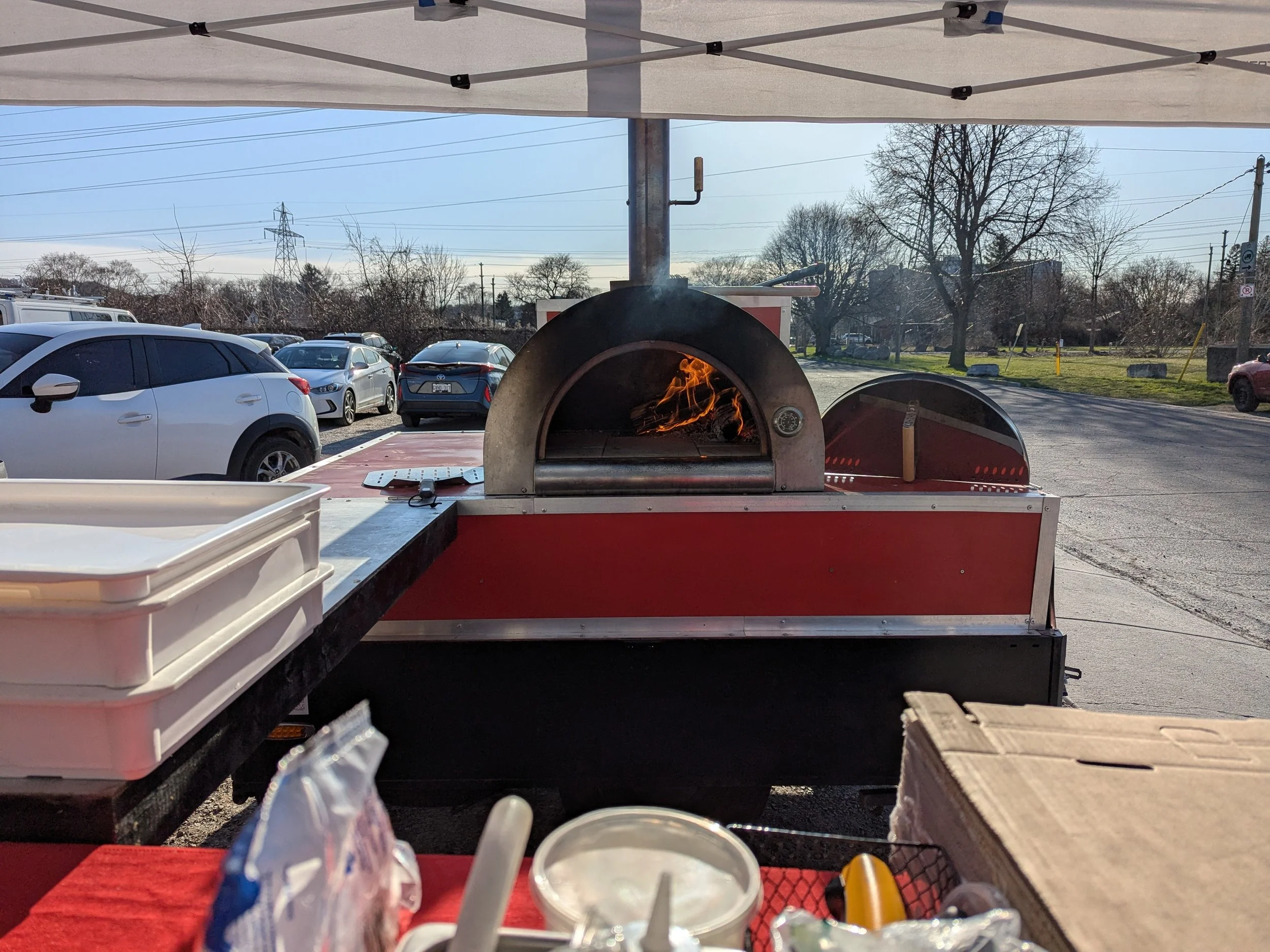 A pizza oven with a flame inside at an outdoor food stand, with parked cars and trees in the background.