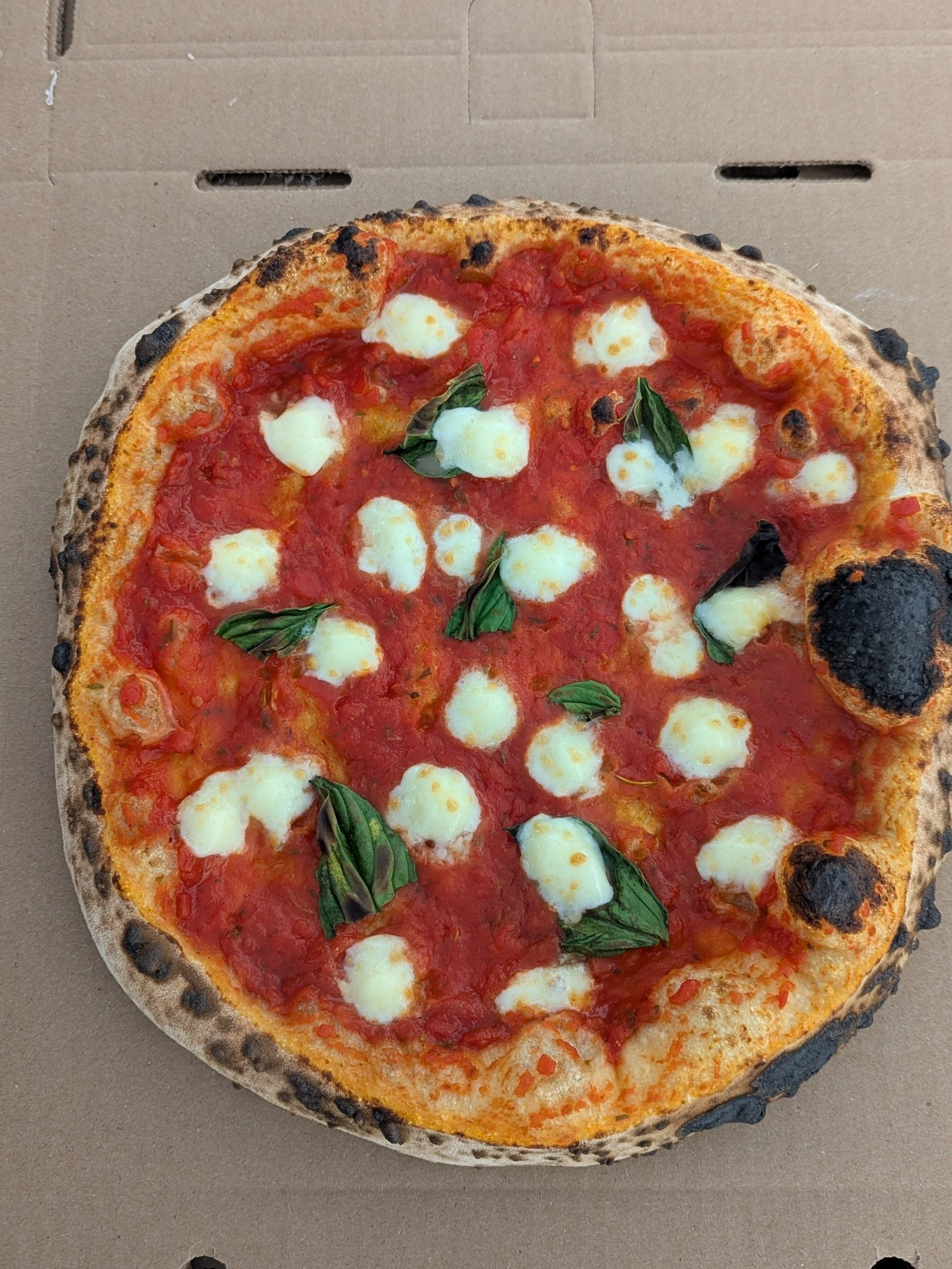 A Margherita pizza with tomato sauce, mozzarella cheese, and fresh basil leaves on top, in a cardboard box.