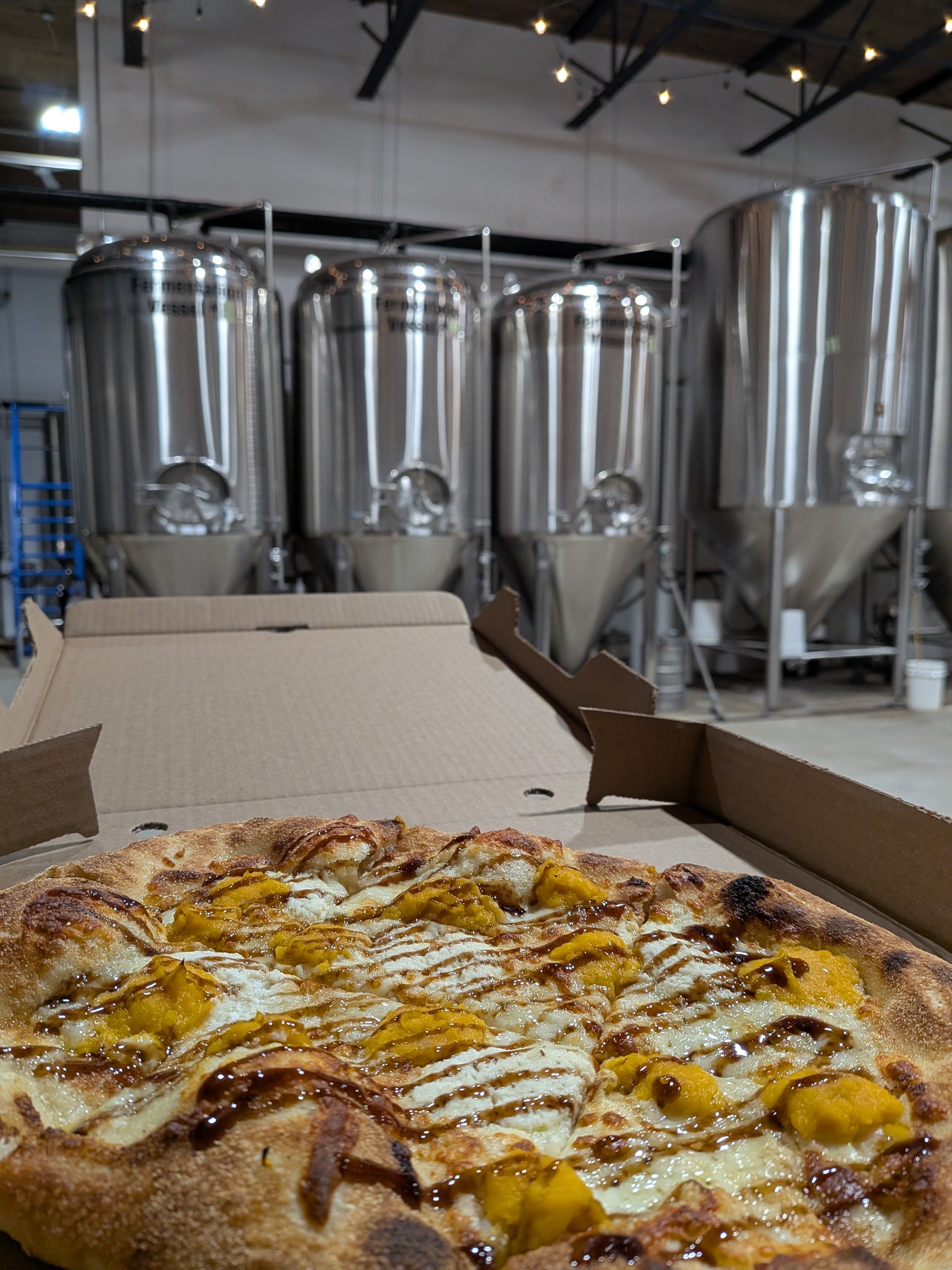 A pizza with yellow toppings, possibly squash or cheese, inside a cardboard box on a table. In the background, industrial stainless steel brewing tanks are visible in a brewery or taproom setting.