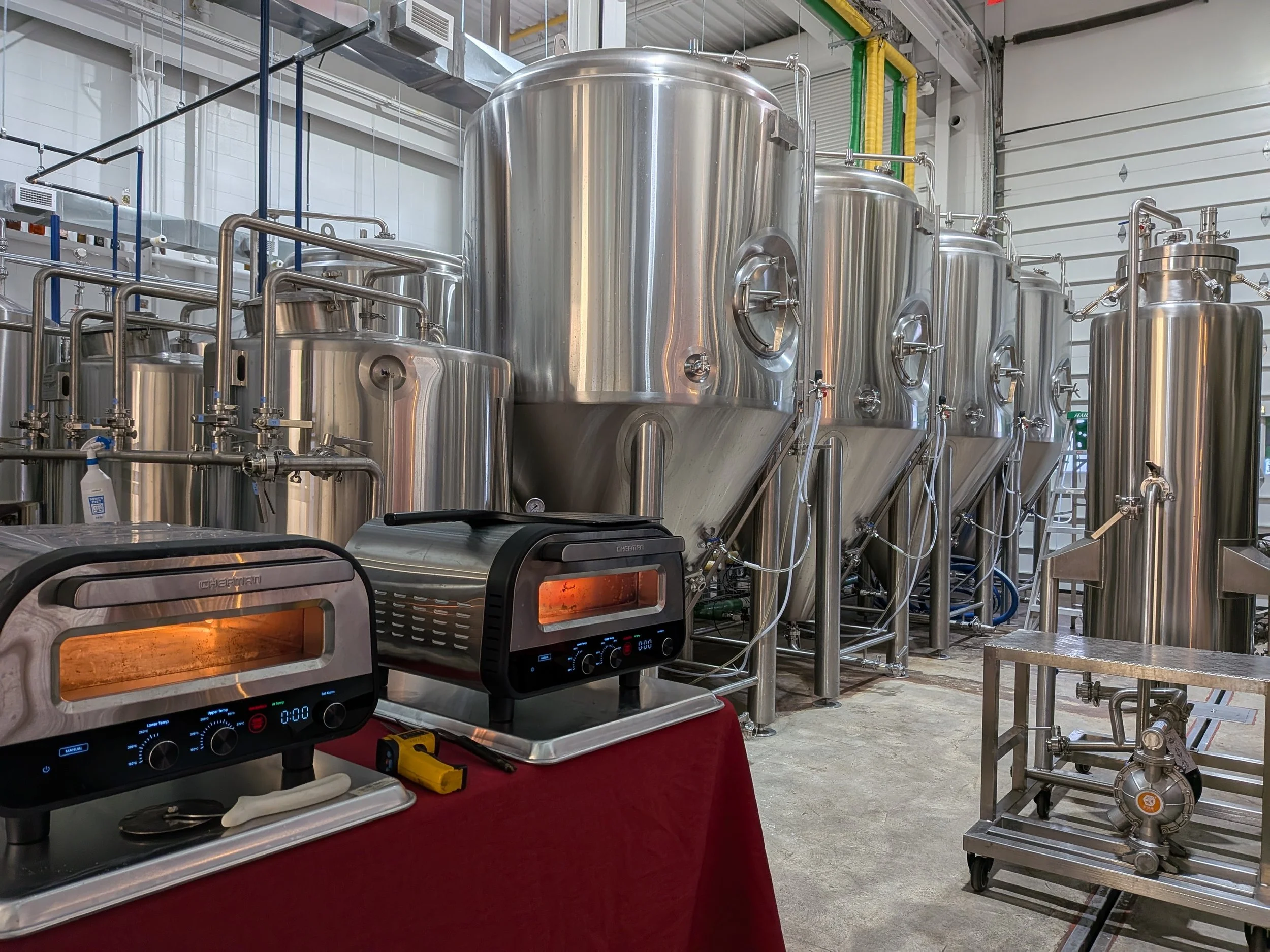 Industrial brewery setup with several large stainless steel fermentation tanks and two small heated ovens on a table in the foreground.