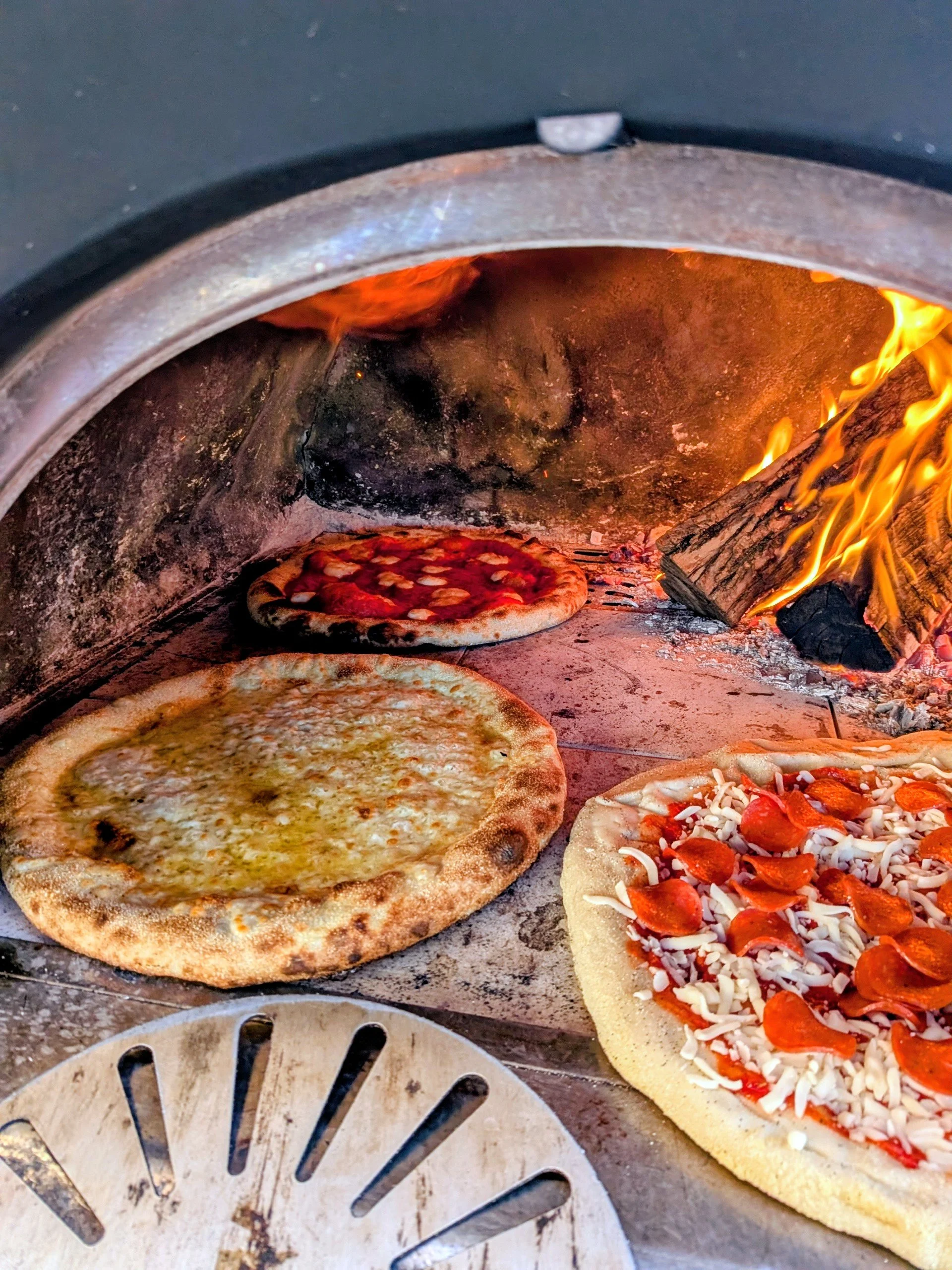 Different types of pizza being baked in a wood-fired oven, with flames visible on the right side.