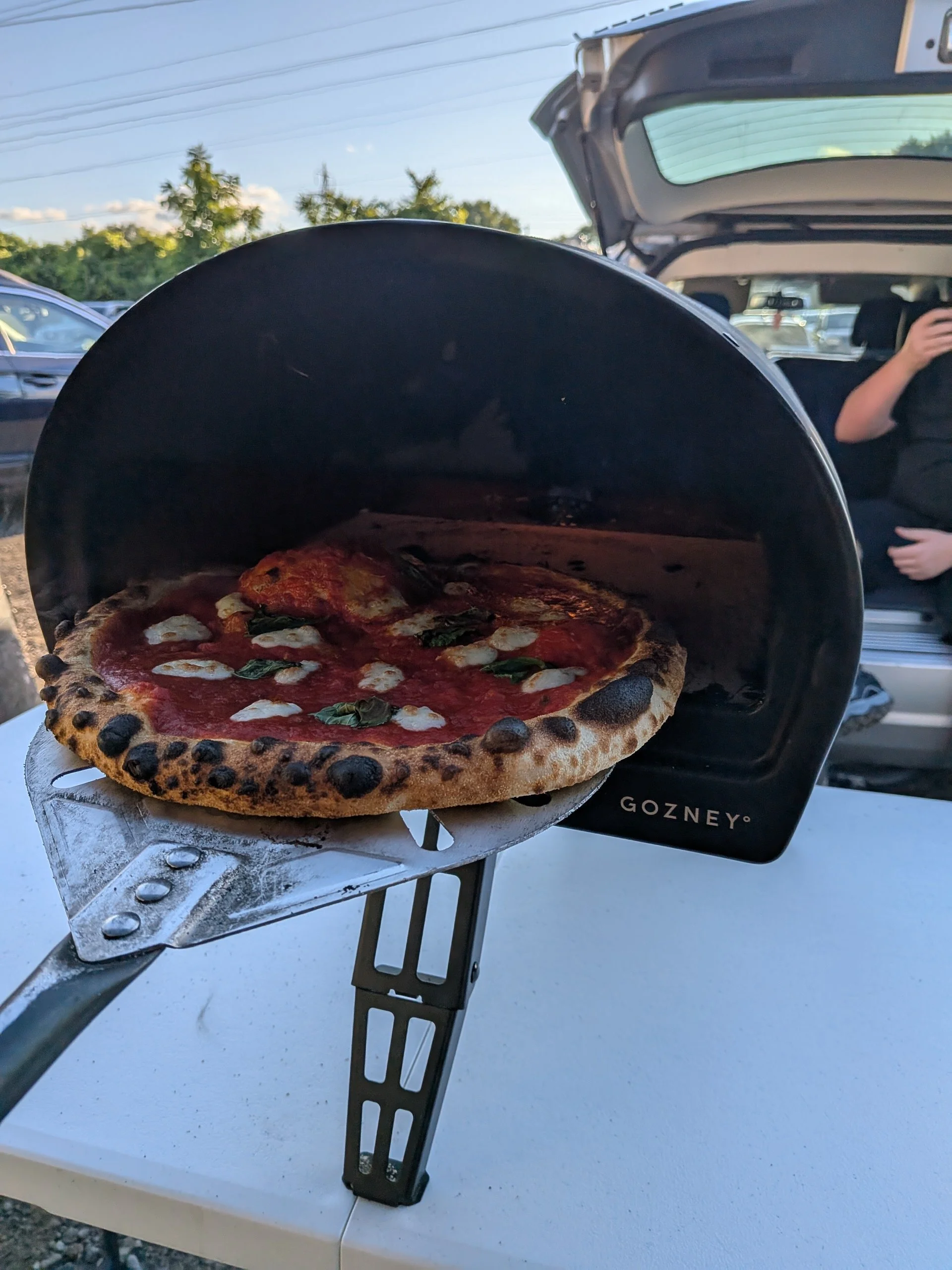 Pizza baking in a portable oven outdoors, with a person sitting in a vehicle in the background.