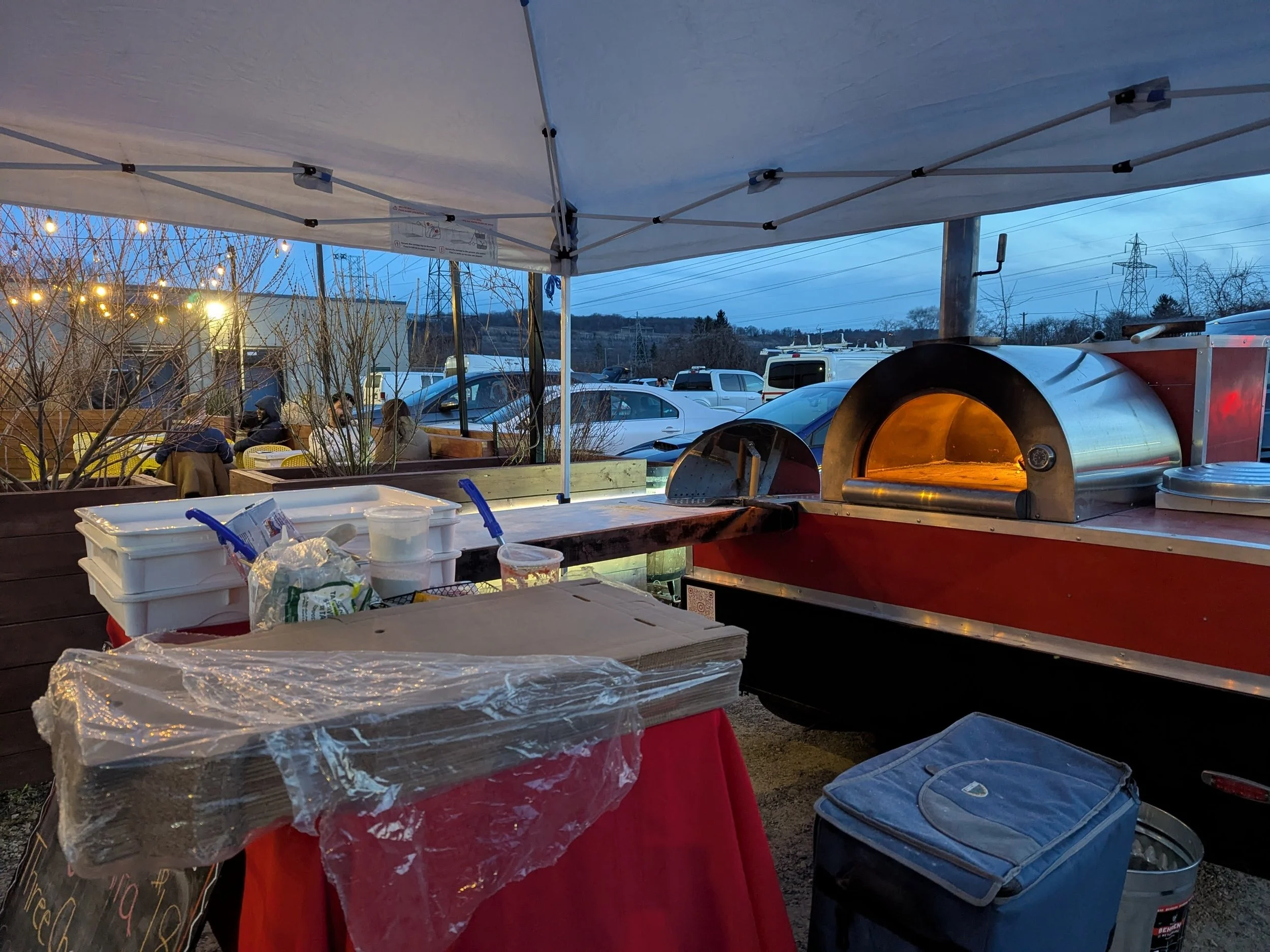 Food stand with pizza oven, covered with a white canopy, outdoor seating area with tables and chairs, and parked cars in the background during evening.