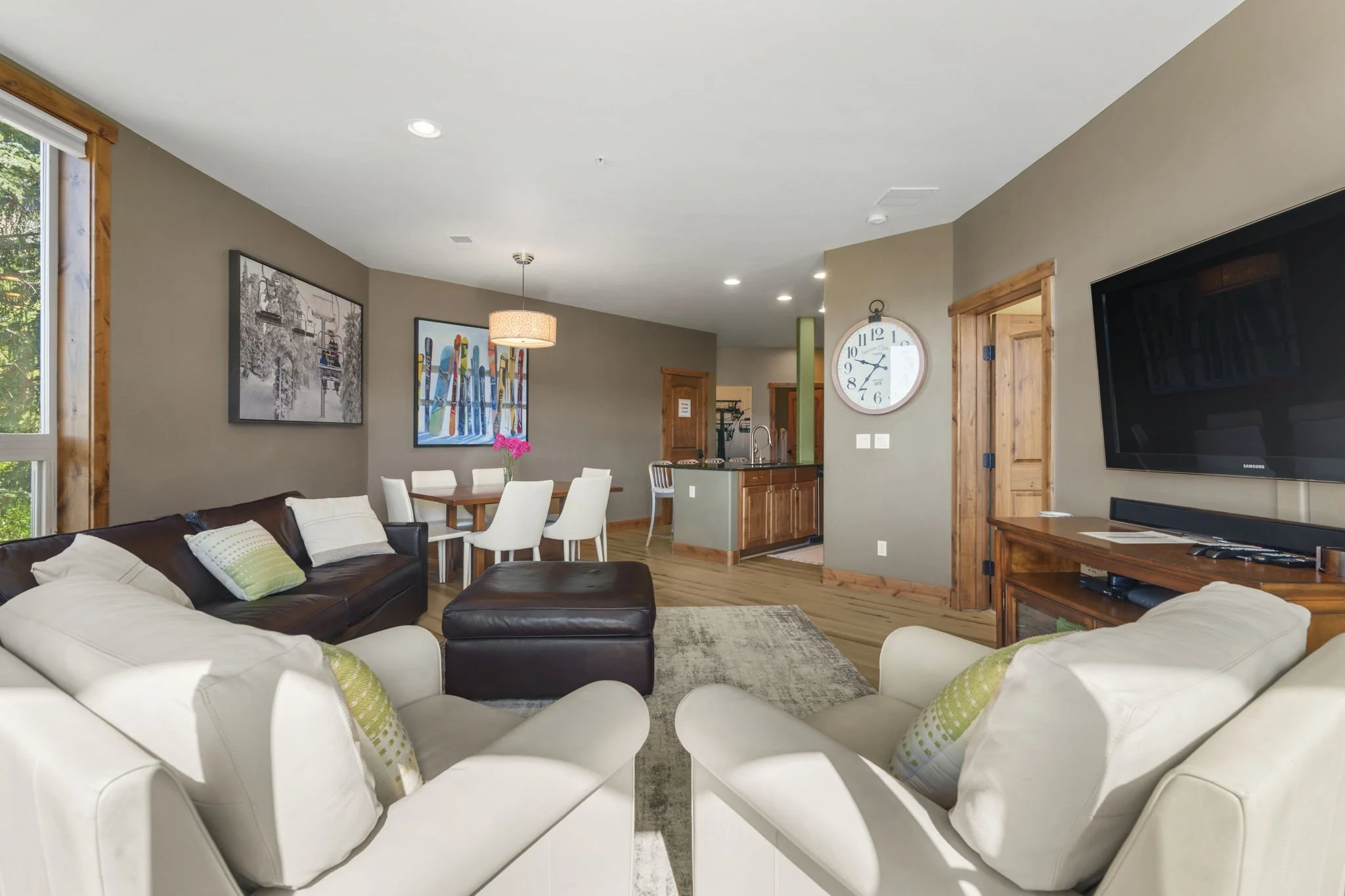 Living room with white couches with lime green pillows, a dark brown leather ottoman, a wooden TV stand, wall art, and a dining area with a table and white chairs.