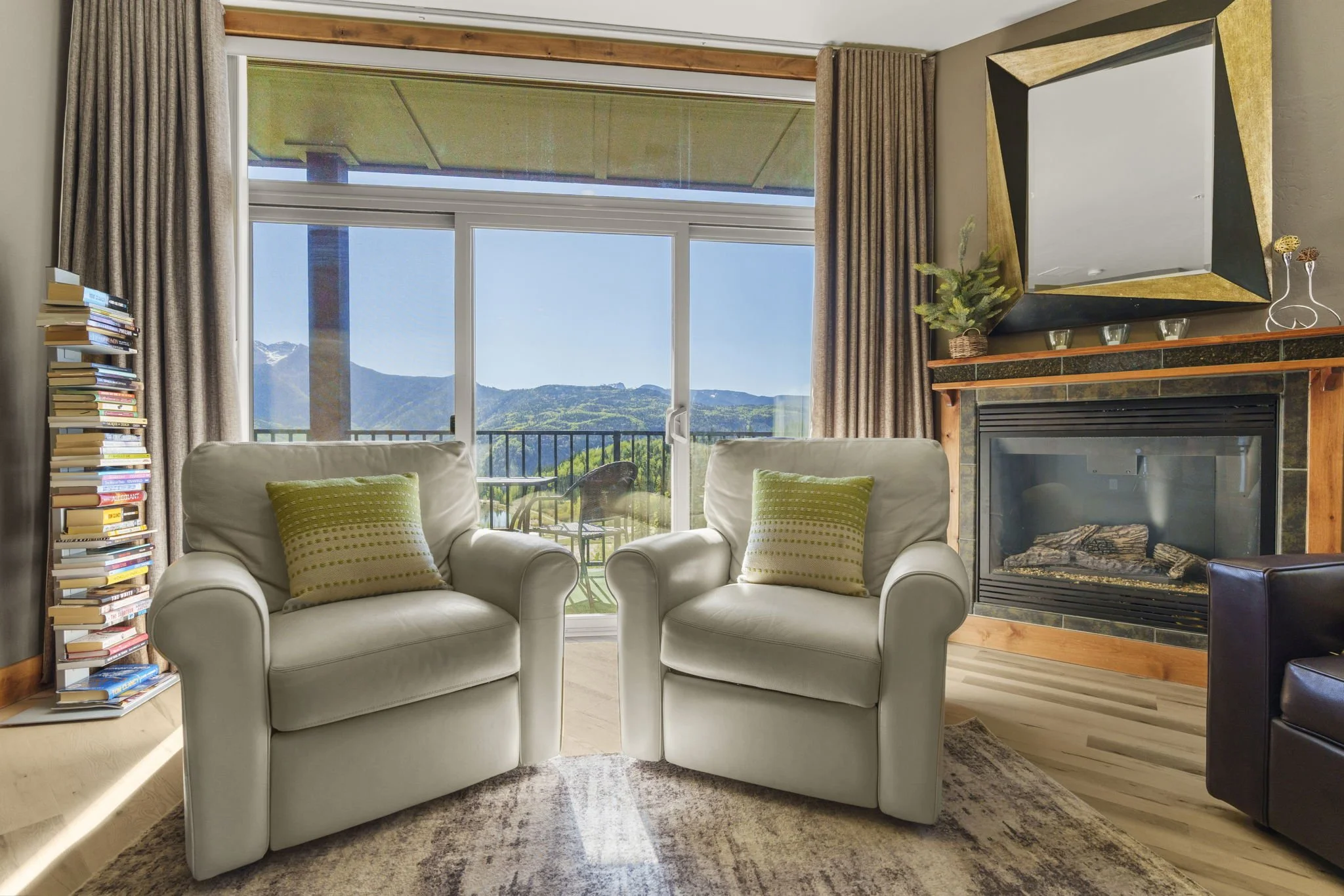 A cozy living room with two white armchairs with green pillows facing a fireplace with a large ornate mirror above it. Large sliding glass doors reveal a mountain view outside, with a balcony and outdoor seating visible.