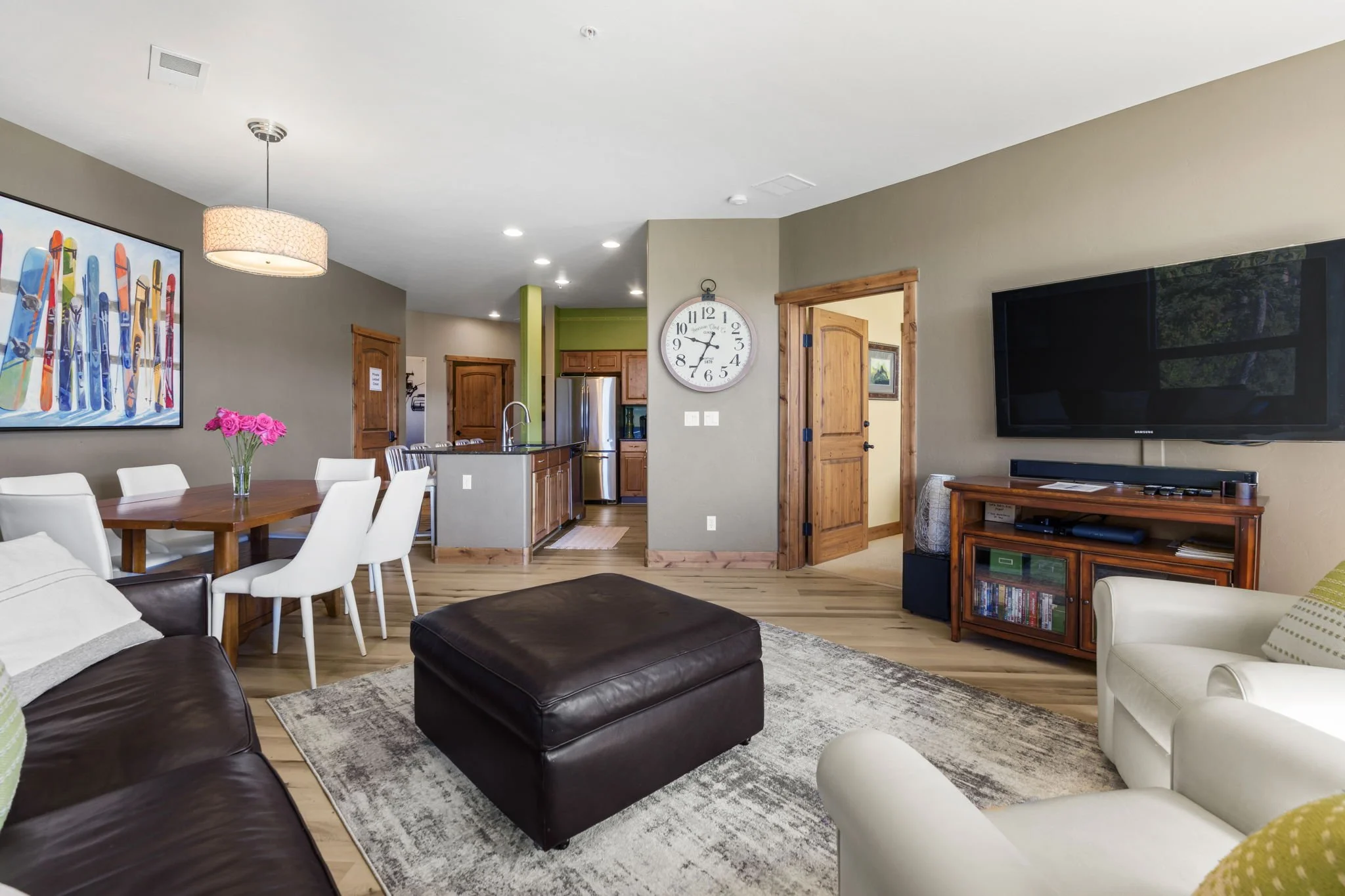 Open-concept living and dining room with a kitchen in the background. Features include a brown leather sofa, a white armchair, a black ottoman, a wooden dining table with white chairs, a large wall clock, a flat-screen TV, and colorful artwork on the