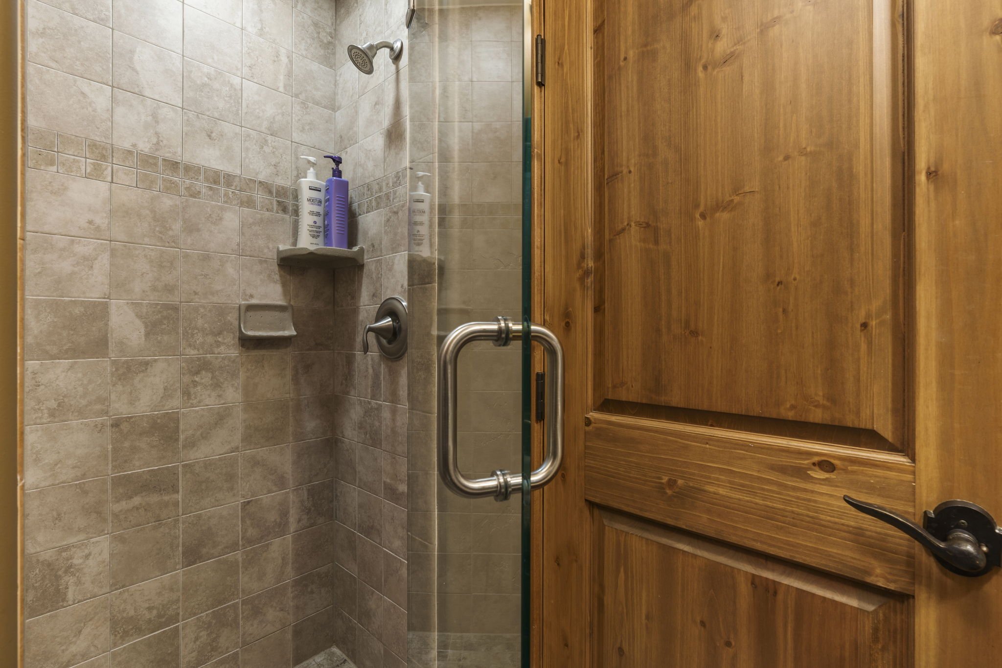 A bathroom shower area with beige tiles, a metal shower head, and a small built-in soap dish. Two bottles of soap or shampoo are on a corner shelf. The shower is enclosed with a glass door with a metal handle, and there is wood paneling on the outsid