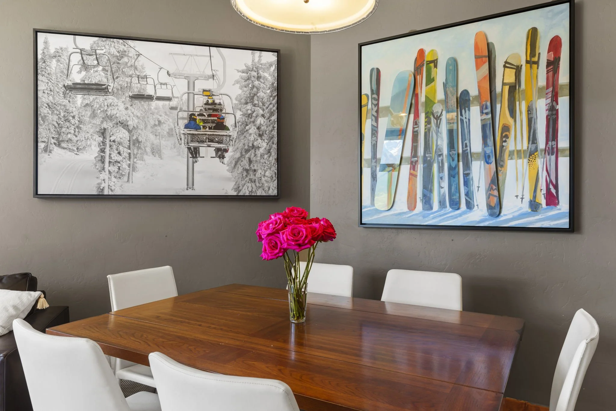 A wooden dining table with white chairs, a glass vase with pink roses, and wall art featuring colorful skis and a black-and-white ski lift photograph in a modern dining room.