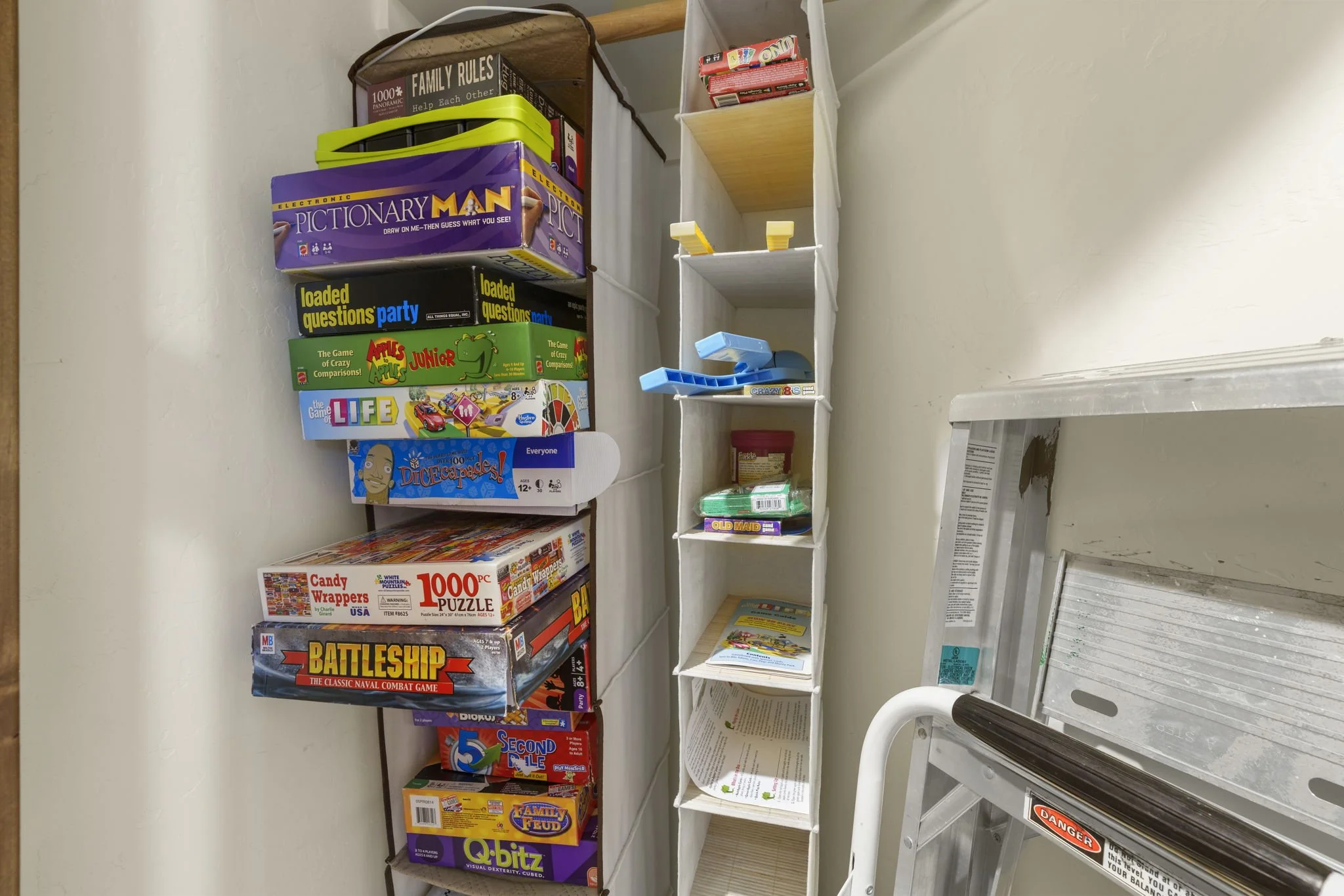 Image shows a room corner with shelves filled with board games and puzzles, including titles like Battleship, Q-bitz, and Apples to Apples, with some empty shelves and a partially visible metal ladder.