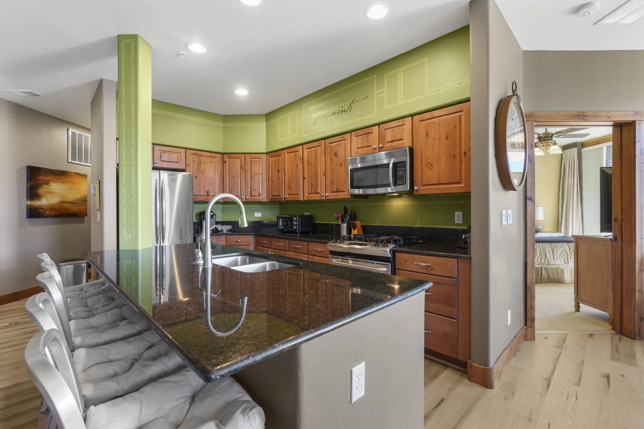 A modern kitchen with wooden cabinets, stainless steel appliances, black granite countertops, a green accent wall with writing, a kitchen island with a sink, and bar stools with gray cushions.