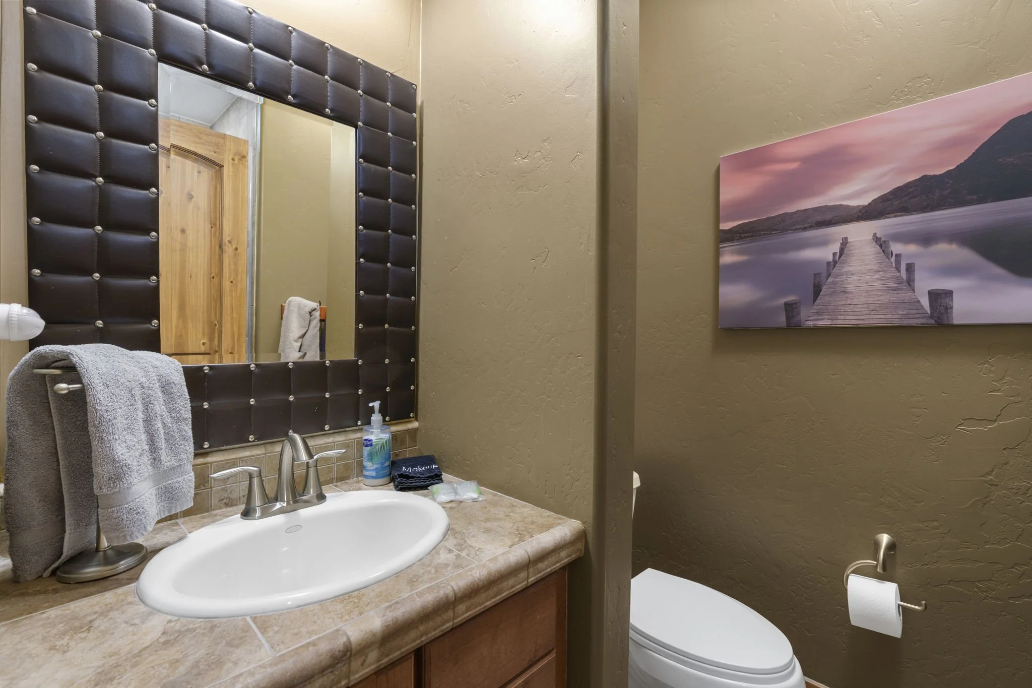 A bathroom countertop with a sink, hand towels, and toiletries, a framed mirror with a tufted leather border, a wall-mounted artwork of a dock extending into a body of water at sunset, and a toilet with a roll of toilet paper.