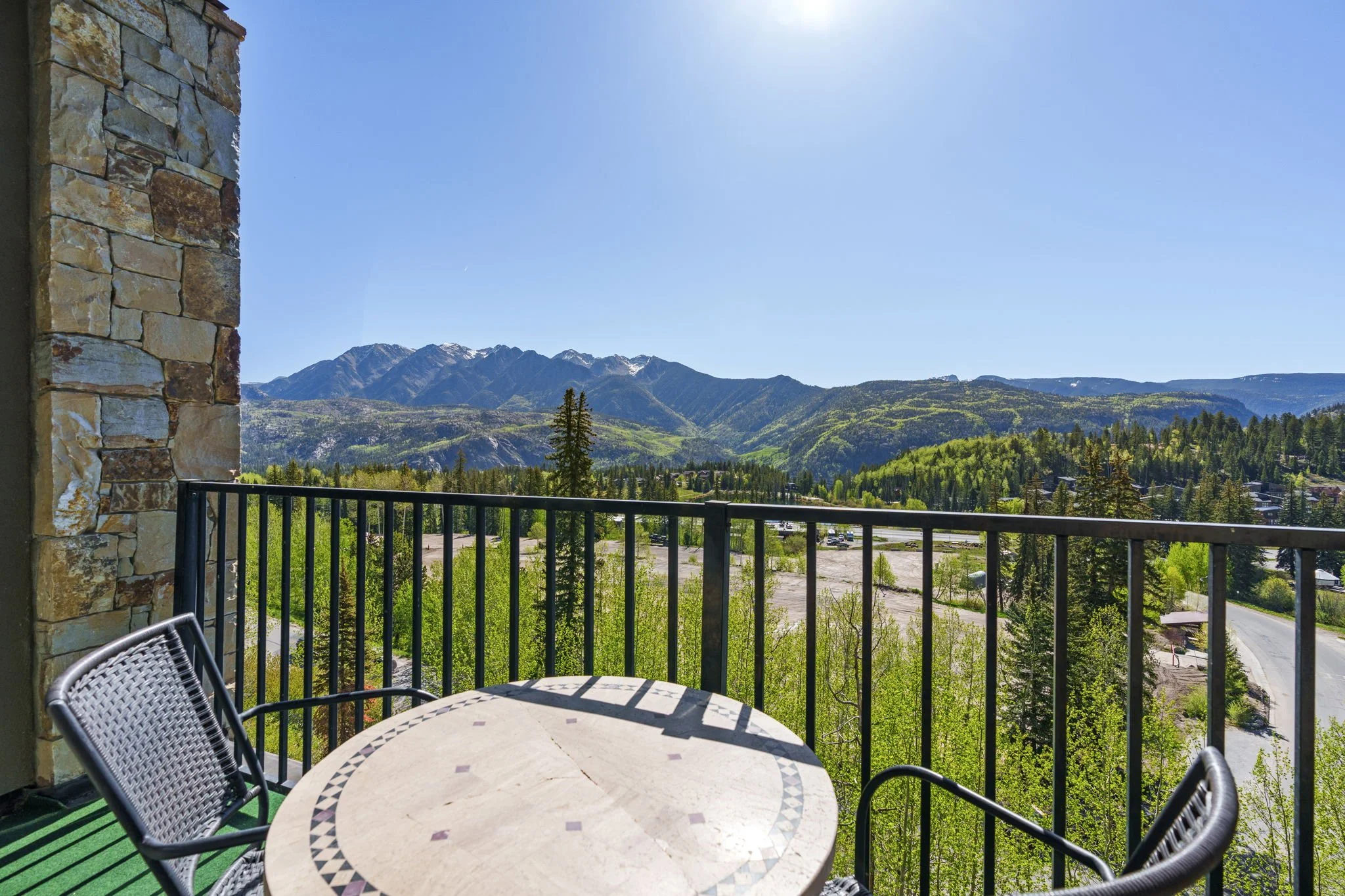 View from a balcony overlooking green hills, mountains with some snow on the peaks, and a clear blue sky.