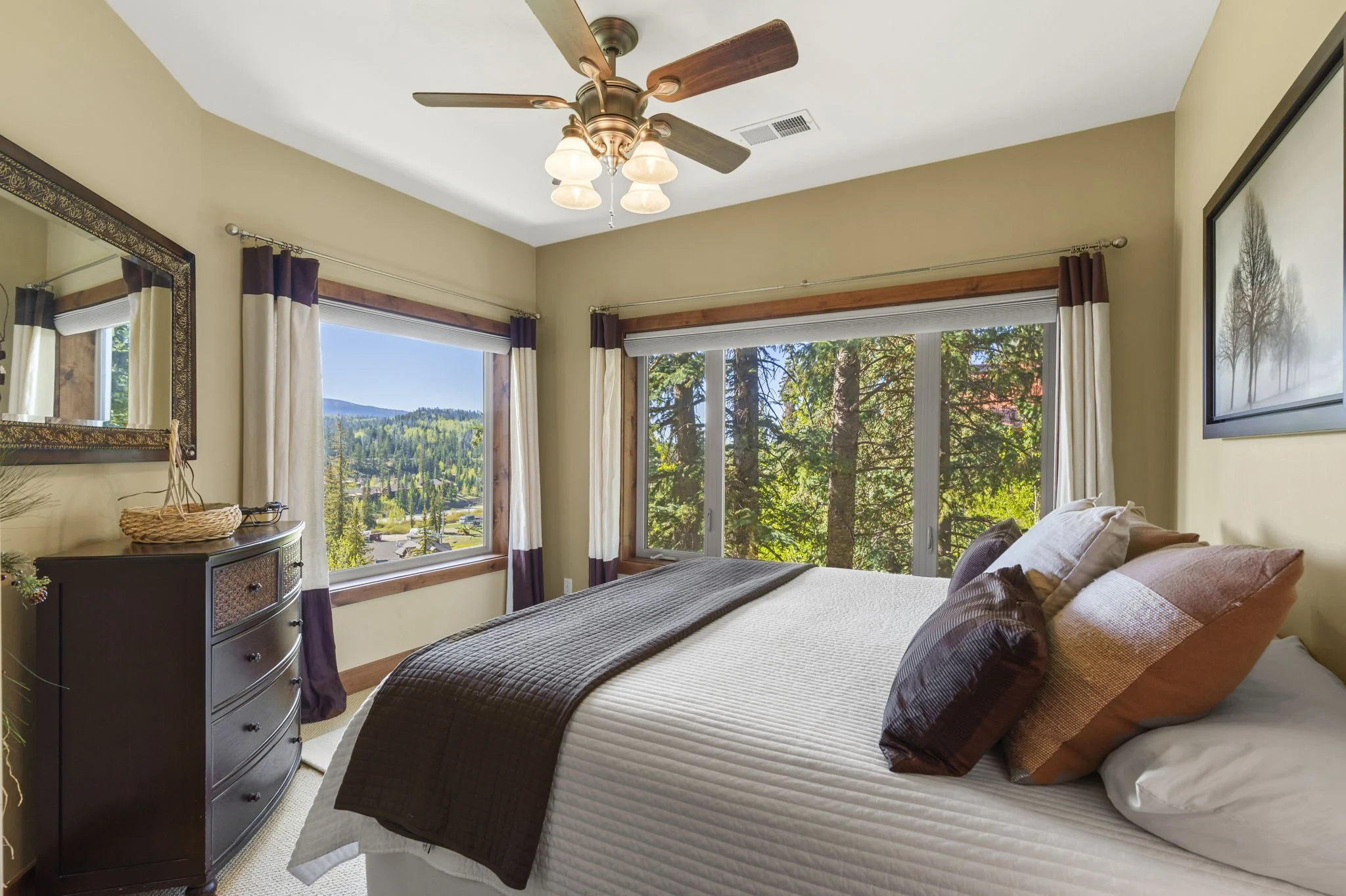 A bedroom with large windows showing a scenic view of trees and hills, a bed with white and brown bedding, a dark wood dresser, and a ceiling fan with lights.