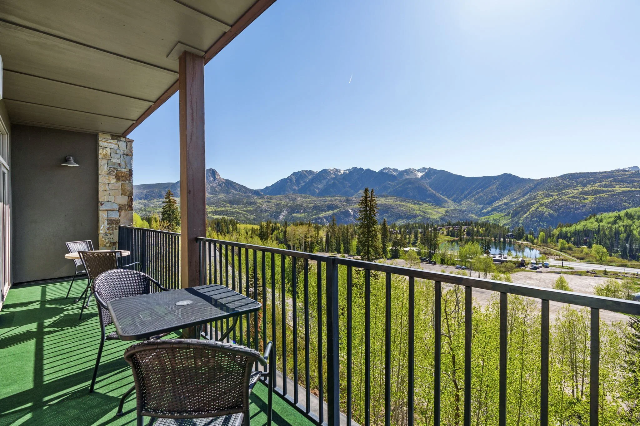 Balcony overlooking green mountains, trees, and a lake with parking lot in the distance, under clear blue sky.