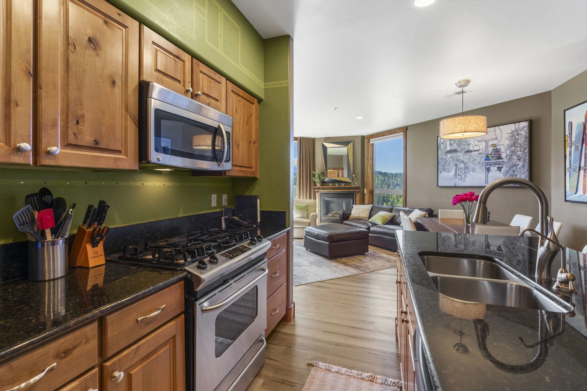 Open-concept living room and kitchen with wooden cabinets, stainless steel microwave, black granite countertops, sink, and cozy seating area with large windows showing a mountain view.