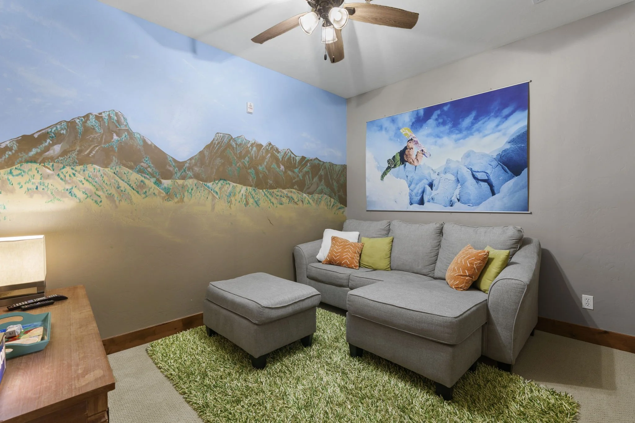 Living room with a gray sectional sofa, colorful pillows, a wooden coffee table, a green shag rug, a large mountain landscape mural on the wall, a ceiling fan with wooden blades, and a window with a curtain.