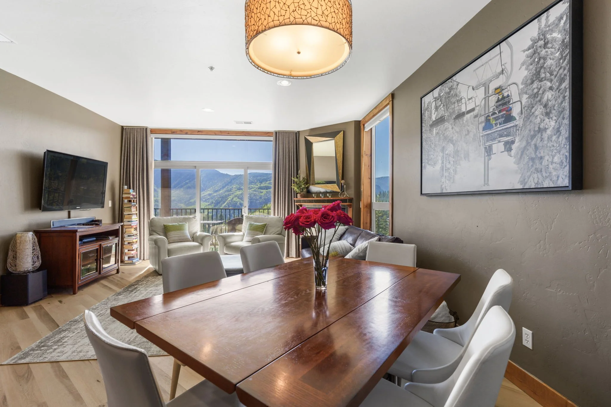 Living and dining area with a wooden dining table, white chairs, a beige sofa with green and beige pillows, a flat-screen TV, a stack of books, a large window showing mountains, a round ceiling light, a framed mirror, a fresh flower vase, and a wall-