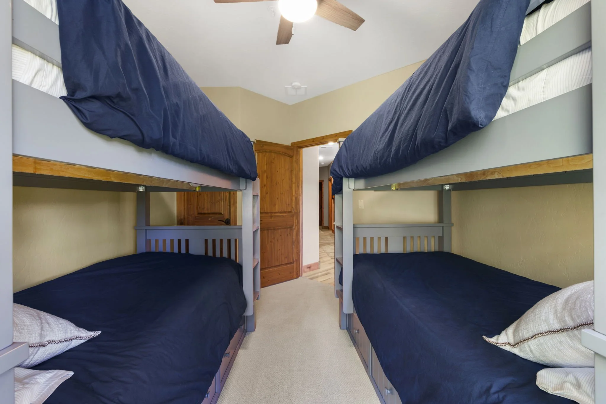 A room with two sets of bunk beds with navy blue bedding, arranged against opposite walls. There is a ceiling fan with wooden blades and a light in the ceiling. The room has light-colored walls and beige carpeting, with a wooden door in the backgroun
