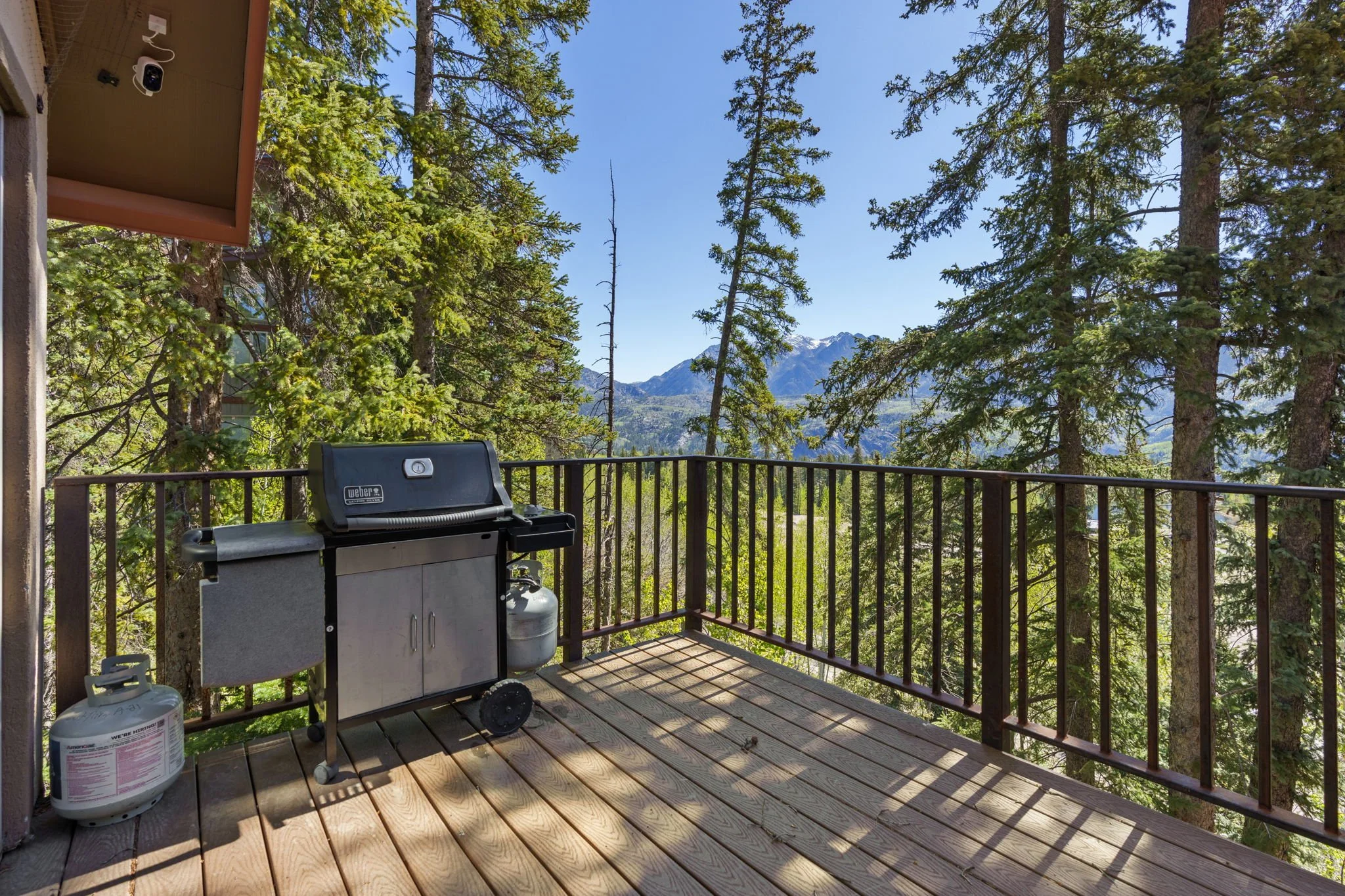 A wooden deck with a barbecue grill, propane tank, and outdoor storage cabinet, surrounded by tall pine trees and mountains in the background under a clear blue sky.
