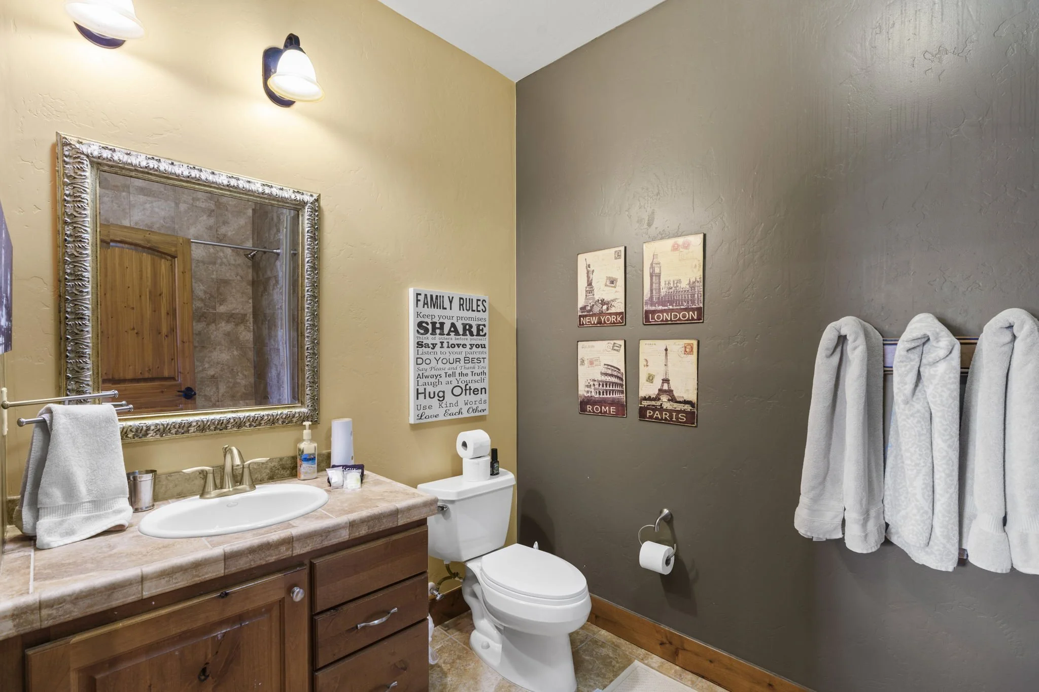 A bathroom with a wooden vanity, a mirror with an ornate silver frame, a sink, a stool with rolled towels, wall art of world landmarks, a family rules sign, and a toilet.