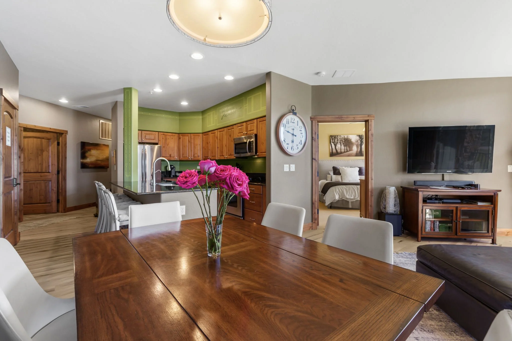 A dining area with a wooden table, white chairs, and a vase of pink roses. In the background, a kitchen with wooden cabinets, stainless steel appliances, and a wall clock. There is a doorway leading to a bedroom with a bed and a framed picture on the