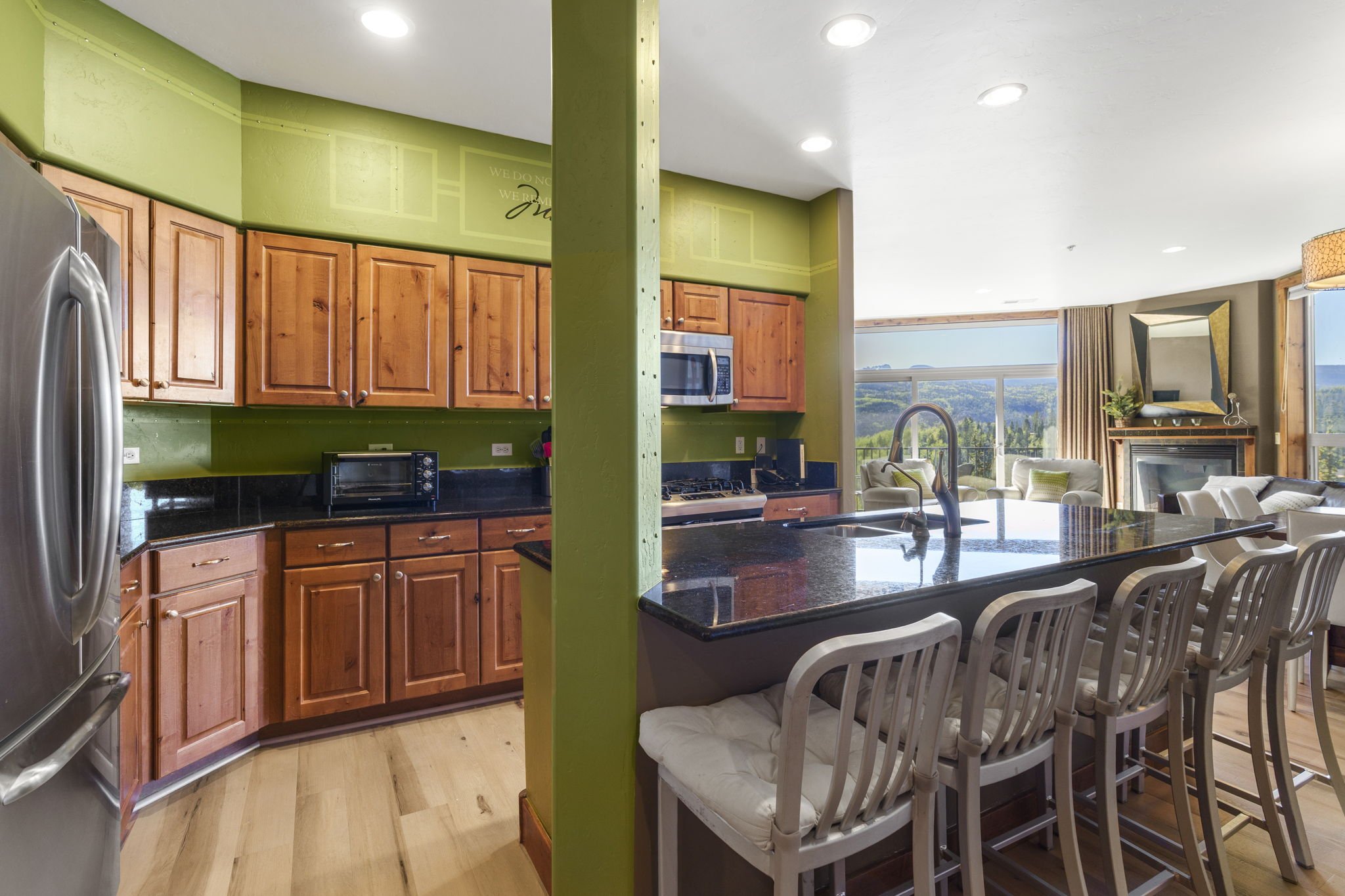 Open-concept kitchen with wooden cabinets, black granite countertops, and stainless steel appliances, adjacent to a living area with large windows showing a scenic view.