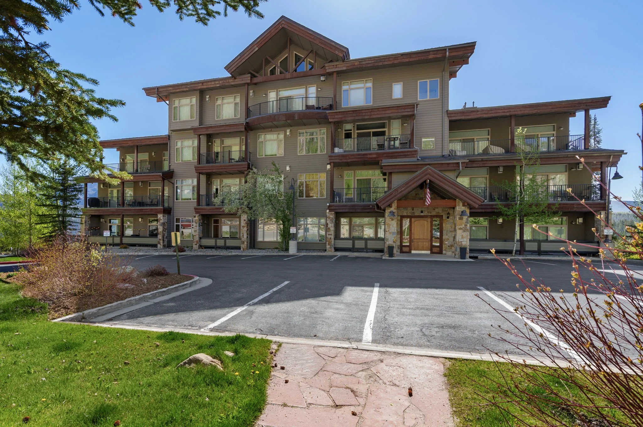 A multi-story residential building with numerous balconies, surrounded by green trees and a parking lot in the foreground.