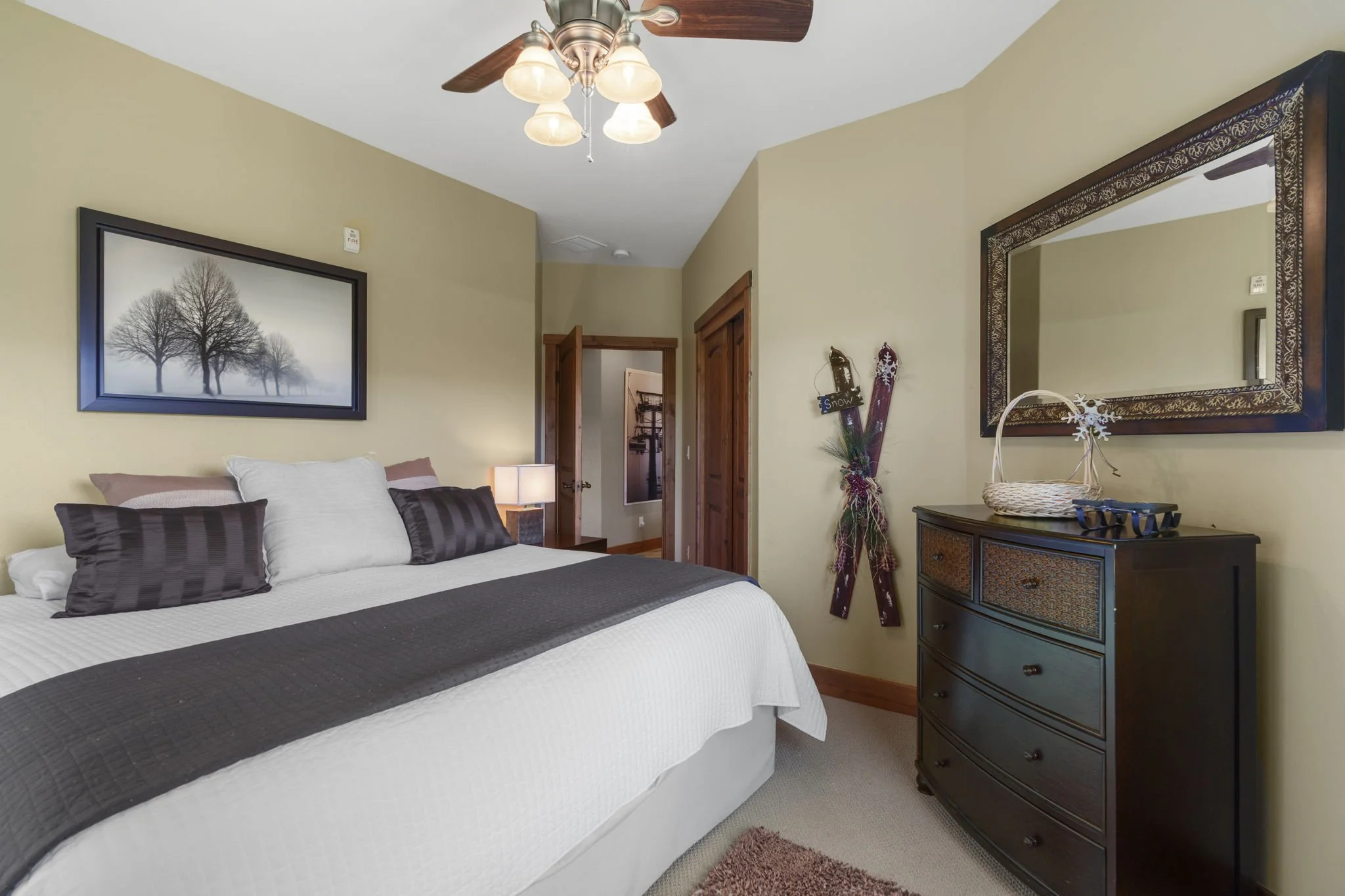 A bedroom with a bed, a wooden dresser with a basket on top, a large mirror, a wall art print of trees, a ceiling fan with lights, and Christmas decorations including snow skis with a small sign and pine branches.