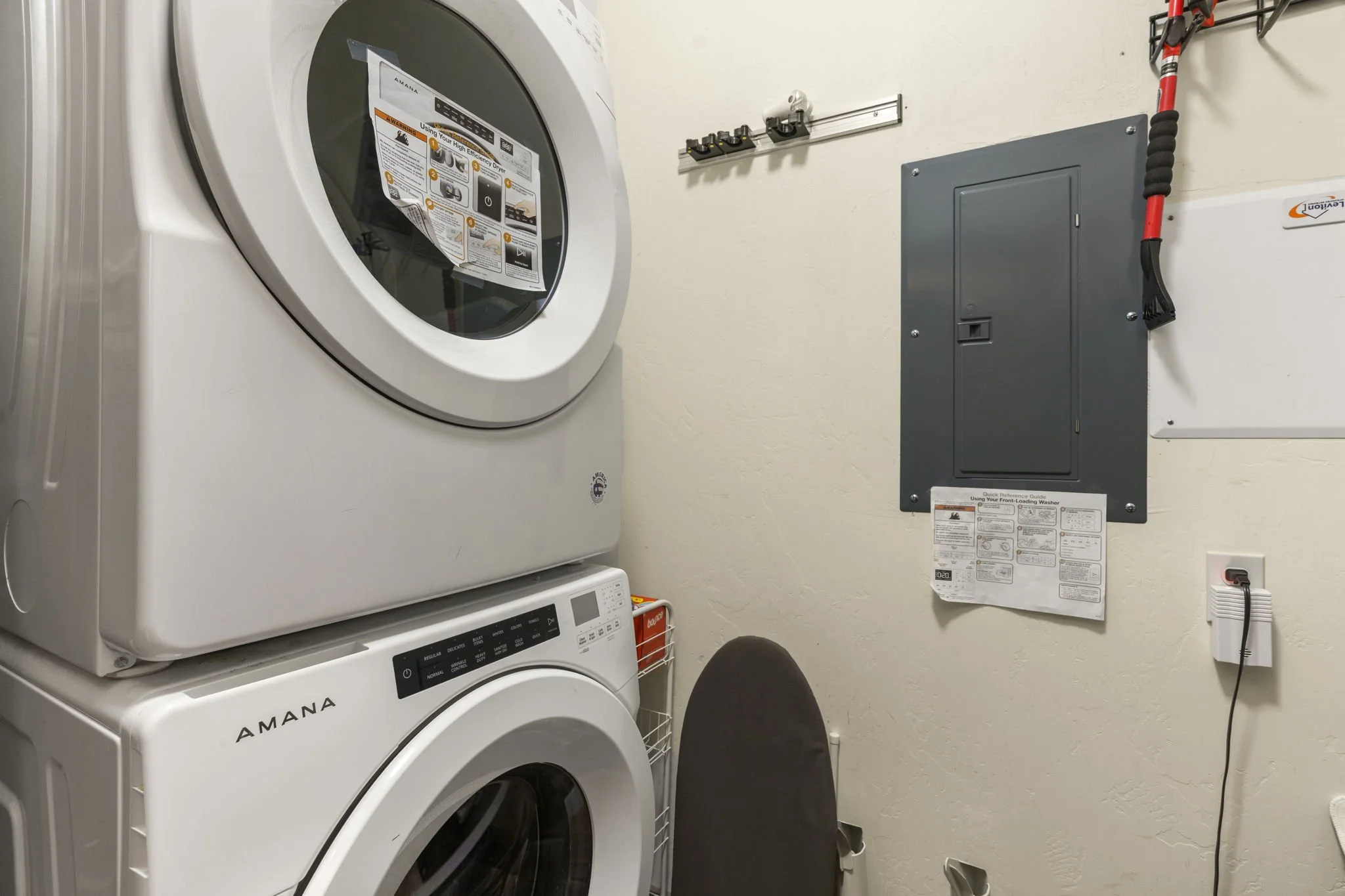 A laundry room with a stacked white Amana washing machine and dryer. There are utility switches, a gray electrical panel, and cleaning supplies on the wall.
