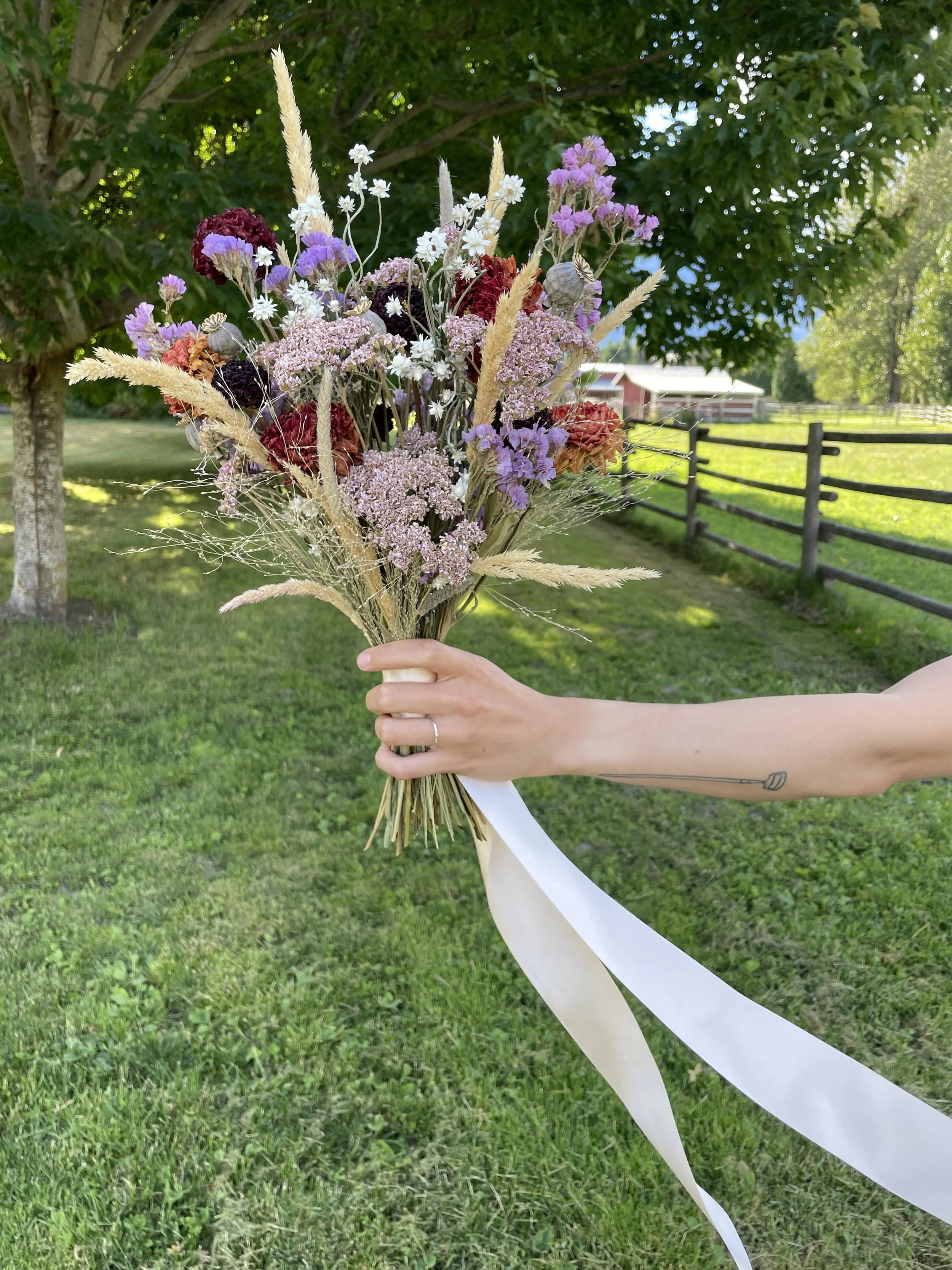 A bouquet of dried flowers, held by a person outdoors with a green, grassy background, trees, a wooden fence, and a barn in the distance.