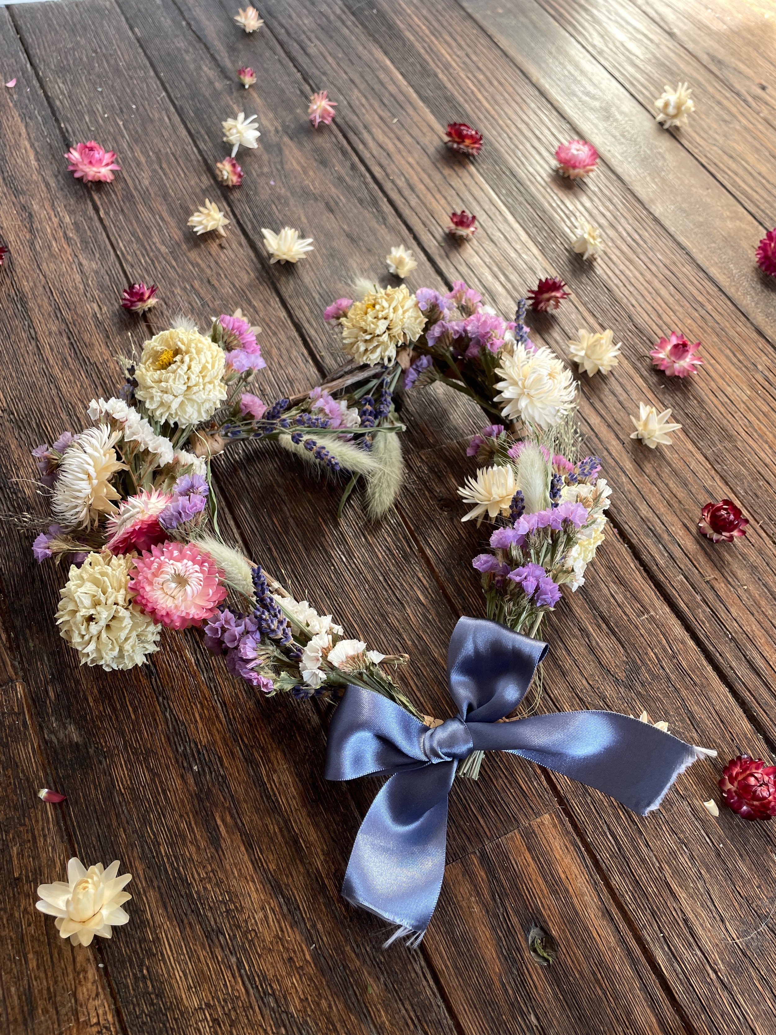 A floral crown made of white, pink, purple, and red flowers with a blue satin ribbon on a wooden surface, with scattered flower petals around it.