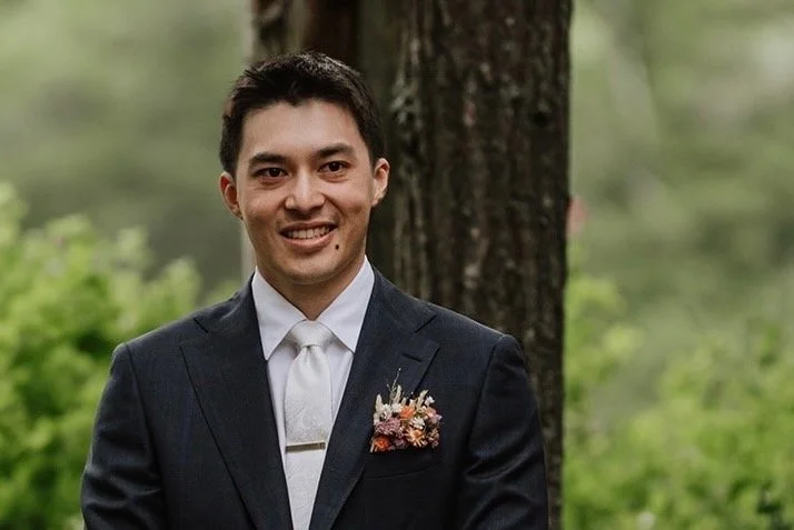 A man in a dark suit with a white shirt and a boutonniere standing outdoors in front of a tree.