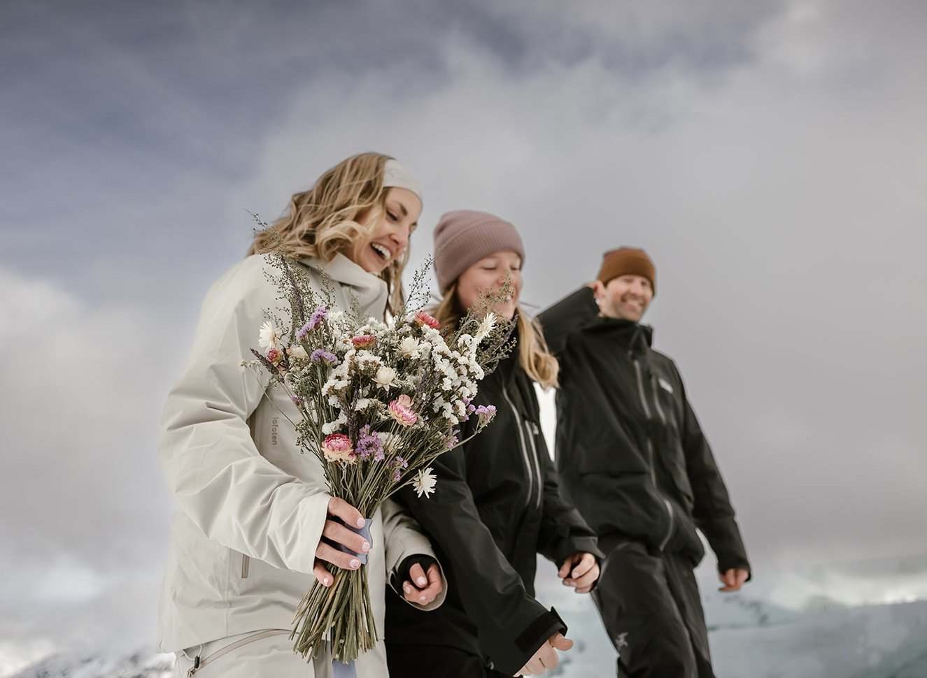 Three people outdoors in winter, holding hands and smiling. The woman in front is holding a large bouquet of flowers.